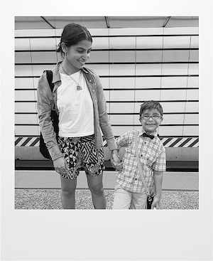 A woman and a young boy stand on a subway platform, holding hands and smiling. The woman wears a jacket, t-shirt, and patterned shorts, while the boy wears glasses and a bow tie. The background shows tiled walls and train tracks.