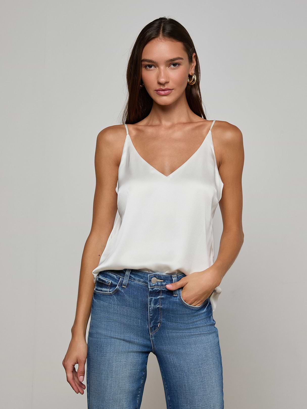 Model wearing the Gabriella Silk Camisole in ivory, paired with blue jeans, stands against a plain light background with one hand in her pocket and a relaxed expression.