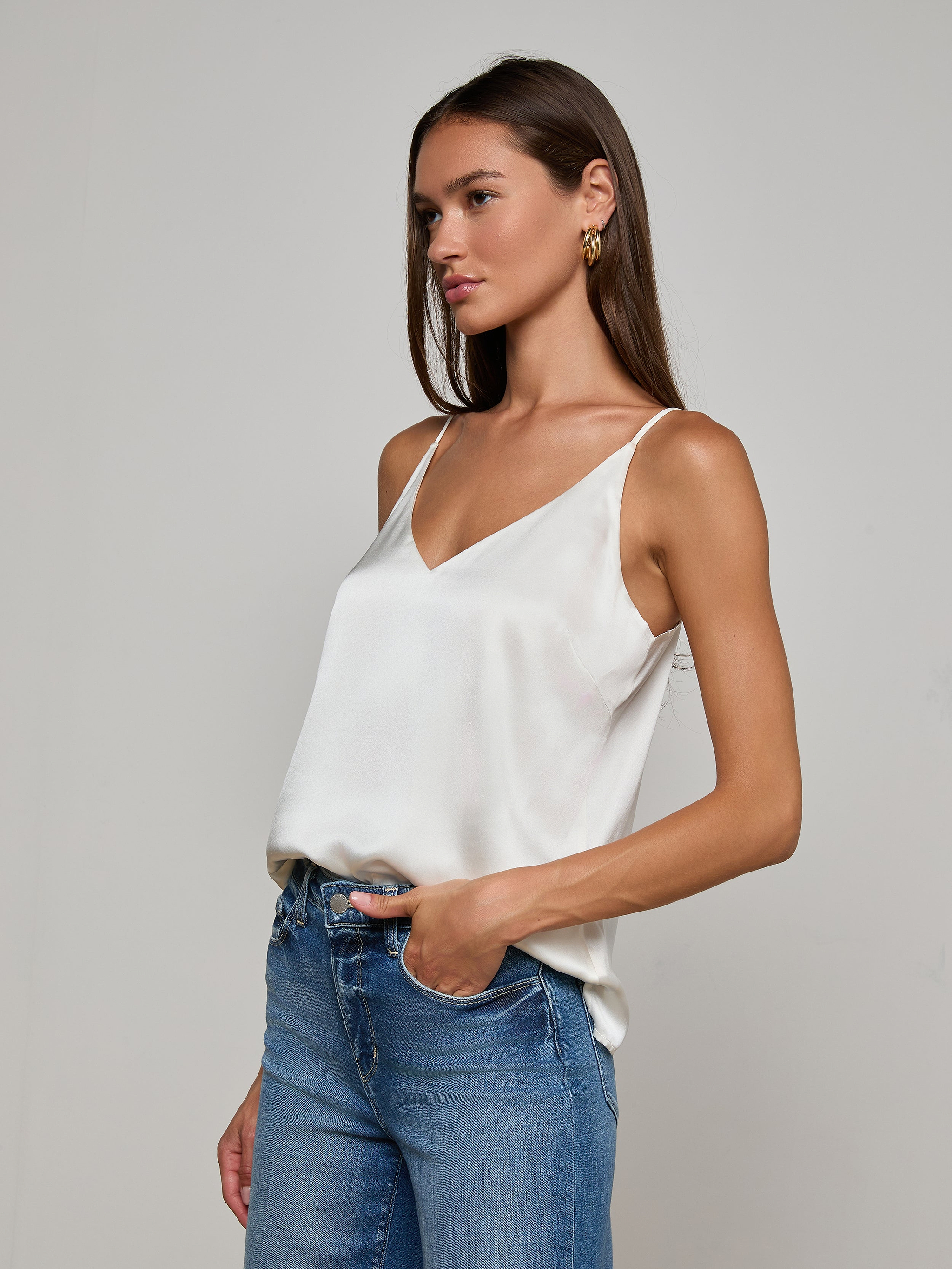 A woman with long brown hair wears the Gabriella Silk Camisole in ivory, paired with blue jeans, standing with one hand in her pocket against a plain light background.