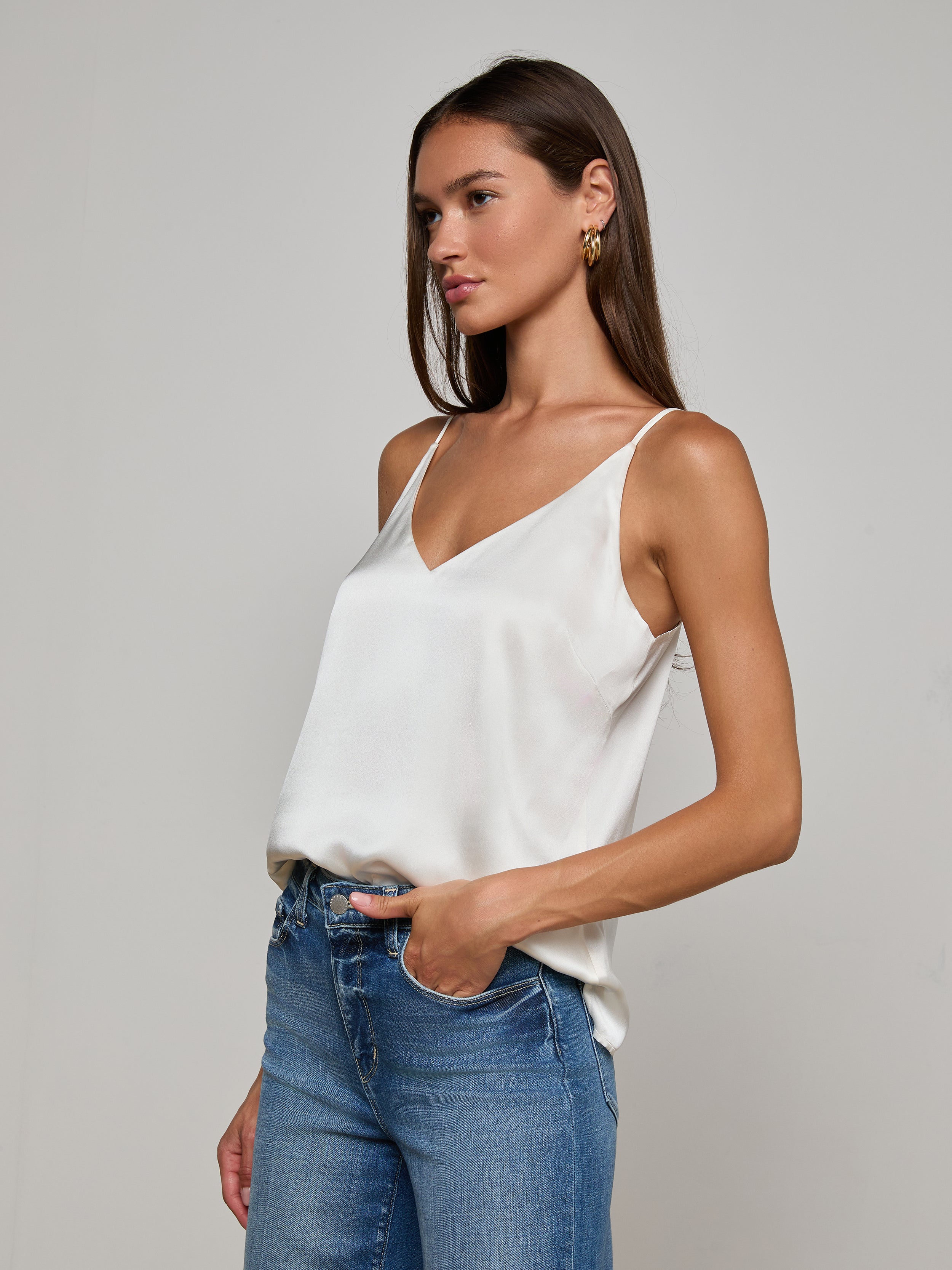 A woman with long brown hair wears the Gabriella Silk Camisole in ivory, paired with blue jeans, standing with one hand in her pocket against a plain light background.