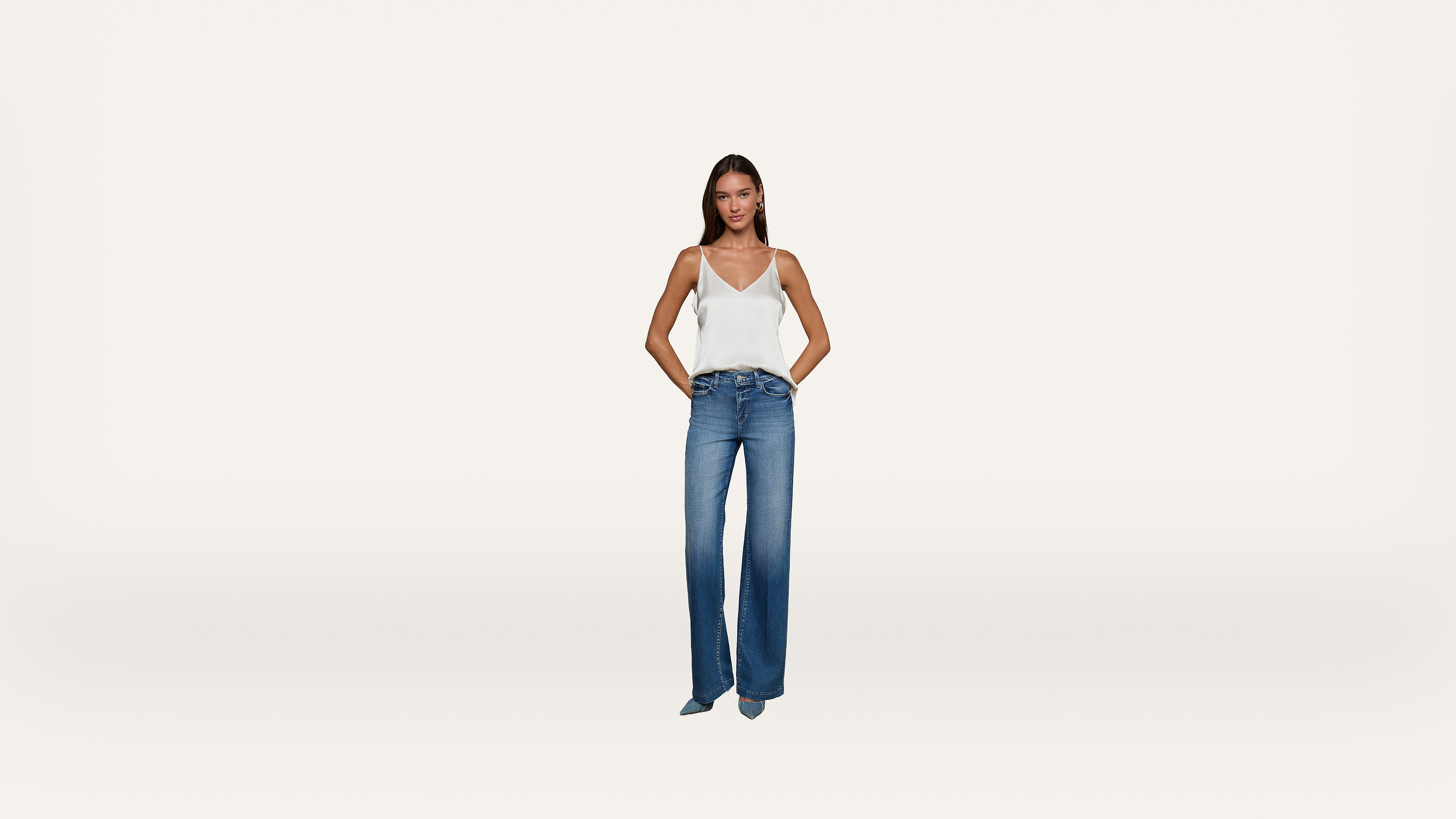 A woman stands with her hands in her pockets, wearing a white sleeveless top and blue jeans against a plain white background.