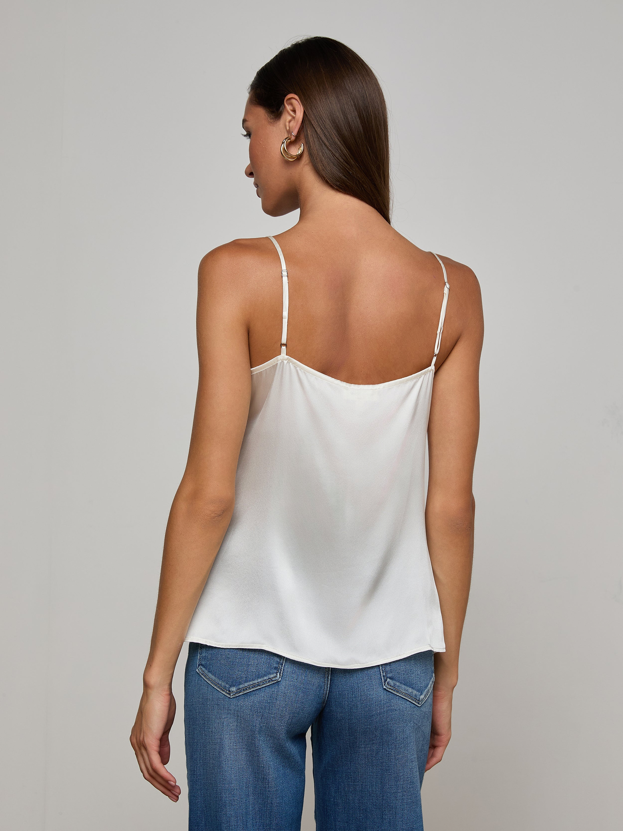 A woman with straight brown hair is facing away, wearing the Gabriella Silk Camisole in ivory with thin straps and blue jeans. The background is plain and light-colored.