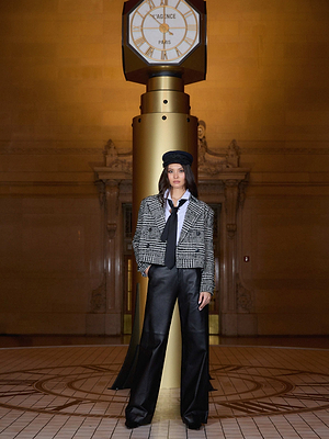 A woman in the Xoey Cropped Trench Coat, white shirt, black tie, and wide-leg black pants stands before a gold clock in a grand, dimly lit hall—perfect for chic fall-to-winter style.