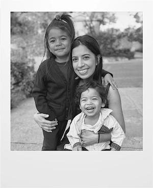 A woman kneels outdoors, smiling and hugging two young children. The older child stands beside her, and the younger child sits on her lap, both smiling. The background shows trees and greenery. The photo is in black and white.