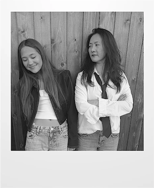 Two women stand side by side in front of a wooden fence. One smiles with eyes closed, wearing a cropped top and jacket; the other looks at her with arms crossed, wearing a shirt, tie, and jeans. Black and white photo.