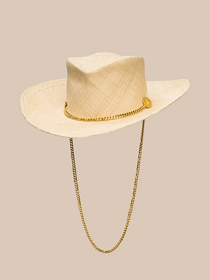 Le chapeau de cow-boy « Zamora » de Gladys Tamez Millinery est un chapeau de cow-boy en paille tressée de couleur crème, fabriqué à la main à Los Angeles. Il se caractérise par un large bord, un ruban en chaîne dorée et une jugulaire, le tout présenté sur un fond beige.