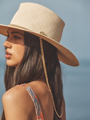 A woman with long brown hair wears the Zamora Cowboy Hat, a woven-straw style featuring a gold chain and L'AGENCE lion head logo, paired with a patterned sleeveless top outdoors against a blurred blue background.