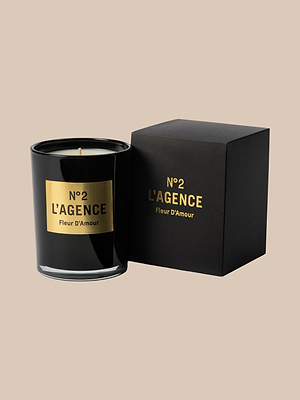 A black hand-poured soy candle labeled Fleur D’Amour in gold text sits beside its matching black and gold box against a plain beige background—a baccarat rose-inspired scent crafted in Southern California.
