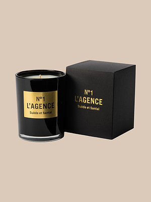 A black candle jar labeled N°1 LAGENCE Suède et Santal in gold lettering sits next to a matching box, both on beige. The luxurious candle features hand-poured soy-blend wax and refined sandalwood notes.