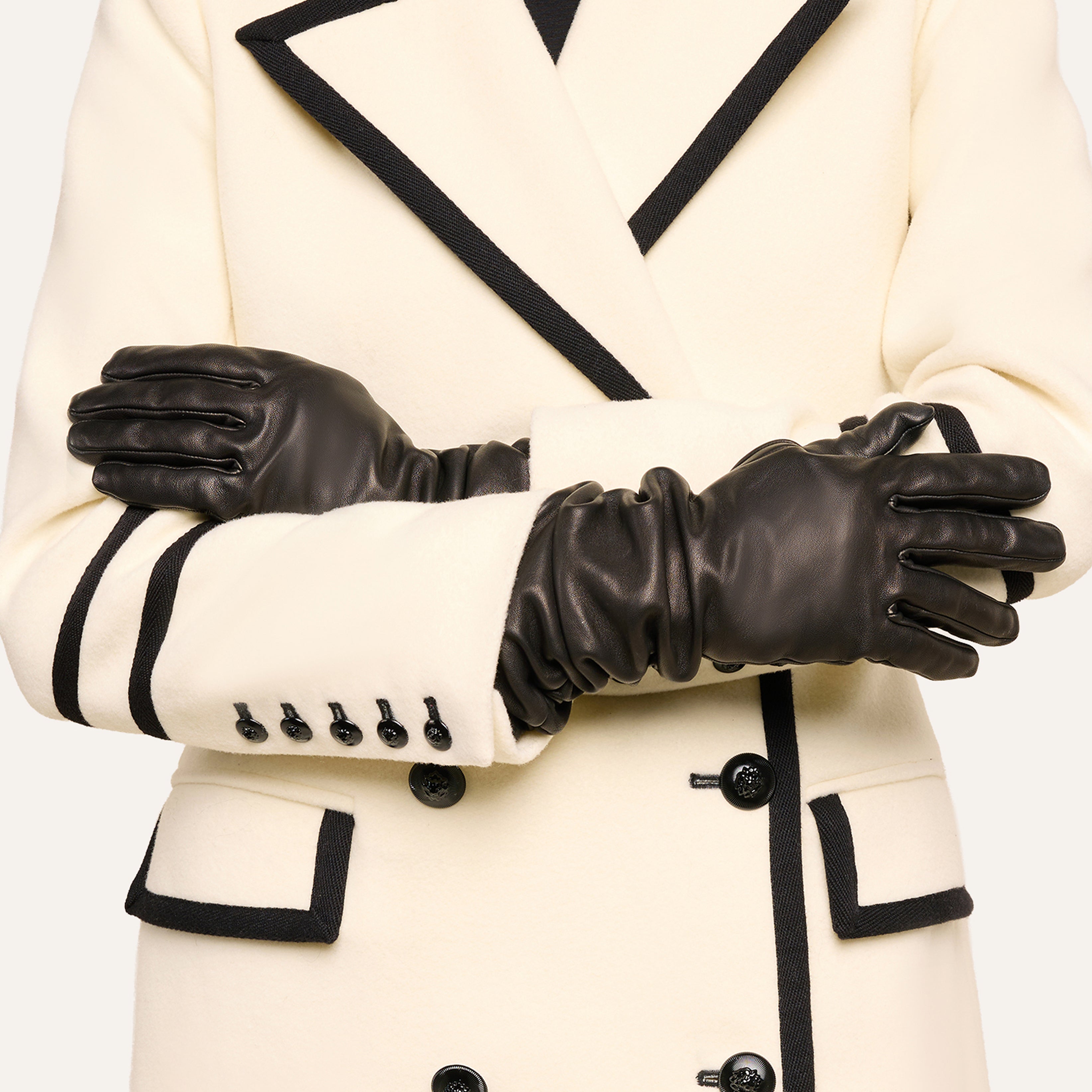 Person wearing a white coat with black trim and black buttons, arms crossed, and wearing black leather gloves. Only the torso and arms are visible; the face is not shown. The sleek black leather gloves add a touch of sophistication to the ensemble.