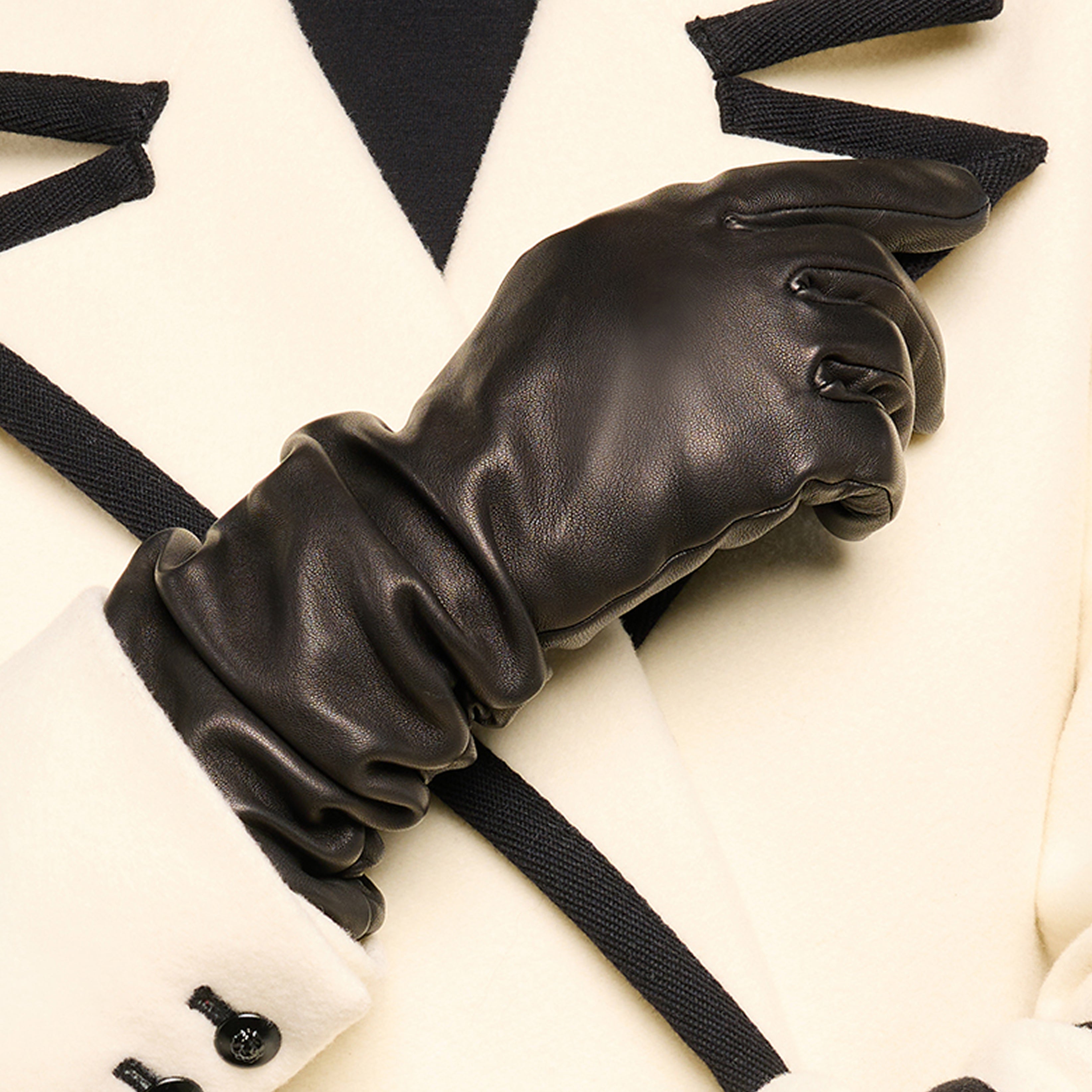 A person wearing a cream-colored coat with black trim stands with their arms crossed, displaying a sleek black leather glove.