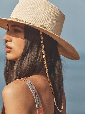 A woman with long brown hair wears a tan wide-brimmed hat with a gold chain and a patterned sleeveless top, standing against a blurred blue background.