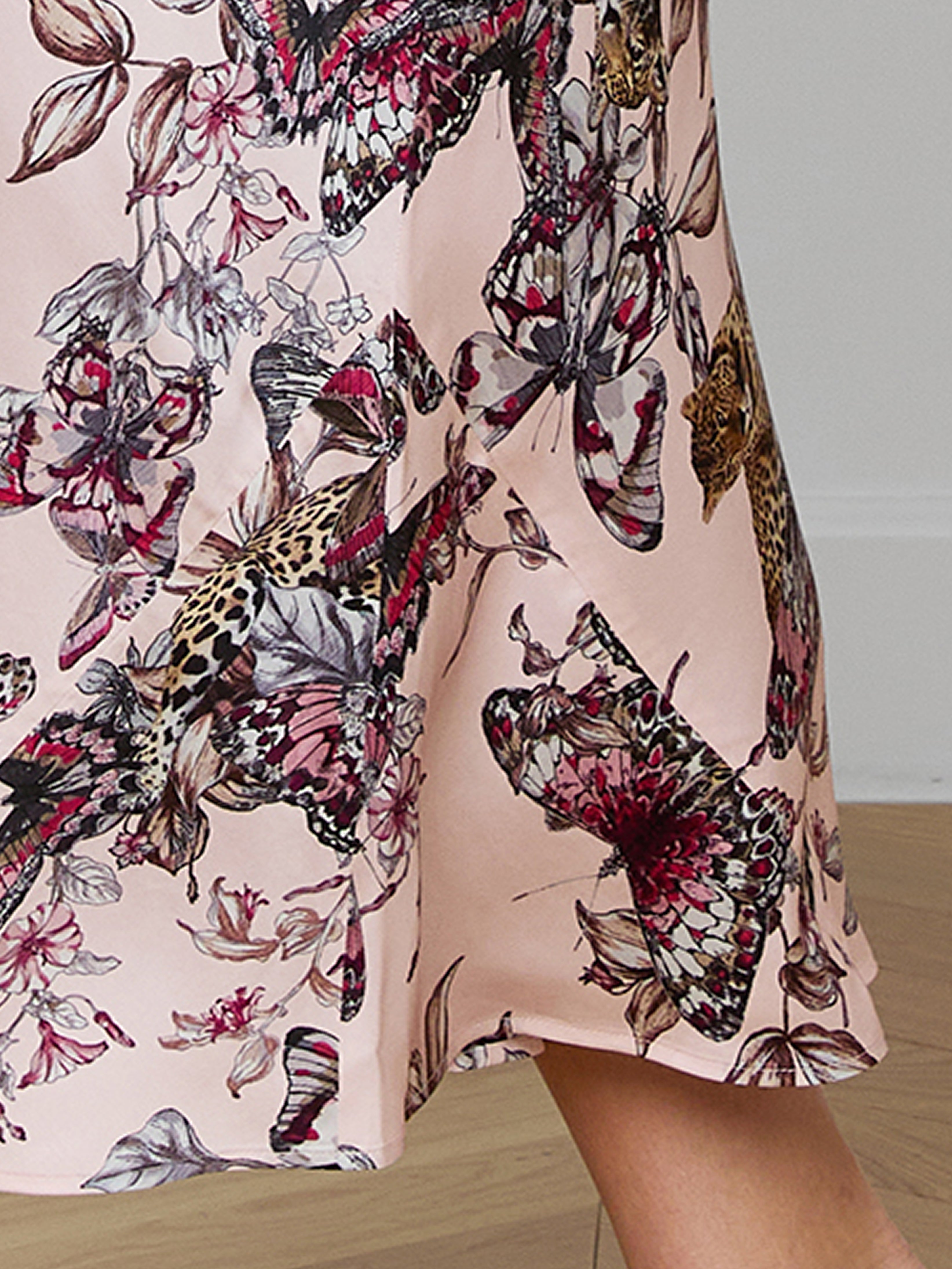 A close-up of a light pink skirt with a colorful butterfly and floral print, featuring shades of black, red, and beige. A persons knee and lower leg are visible at the bottom right of the image.