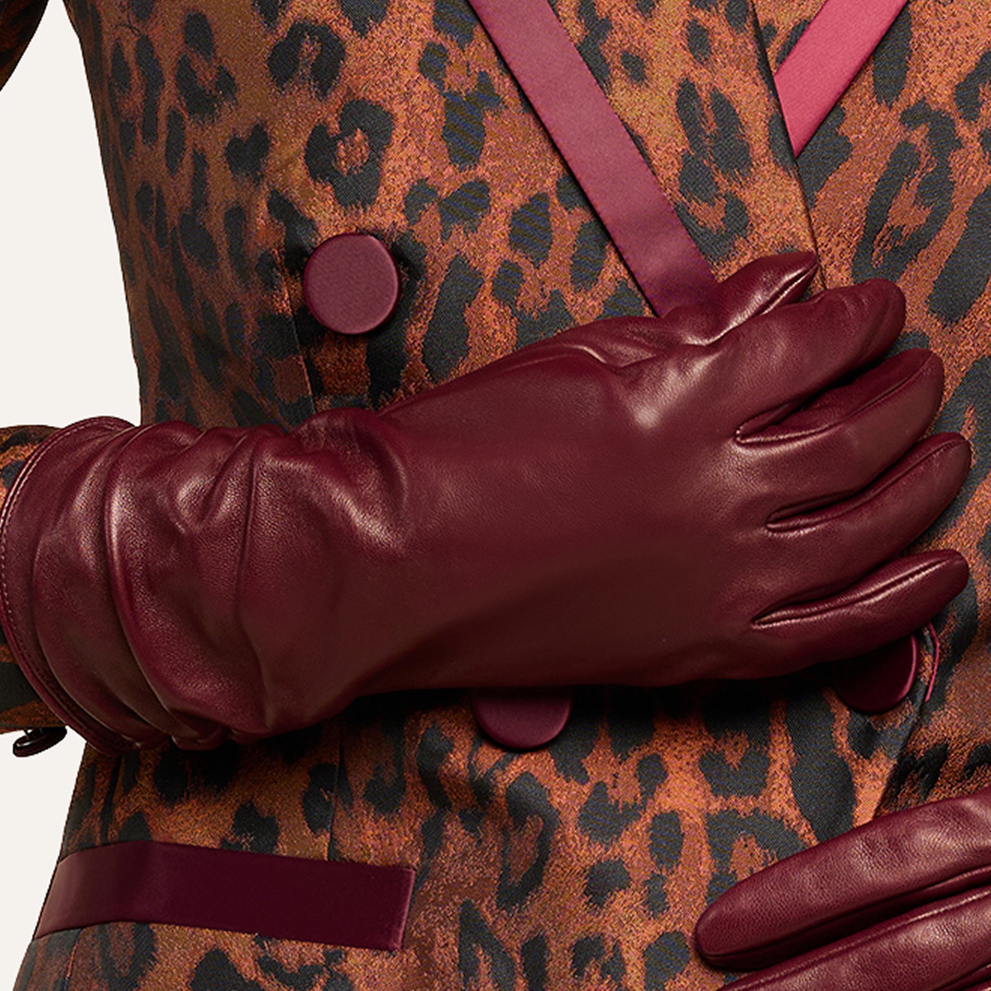 A person wearing burgundy leather gloves holds their hand over a luxurious leopard print jacket adorned with matching burgundy buttons and trim.