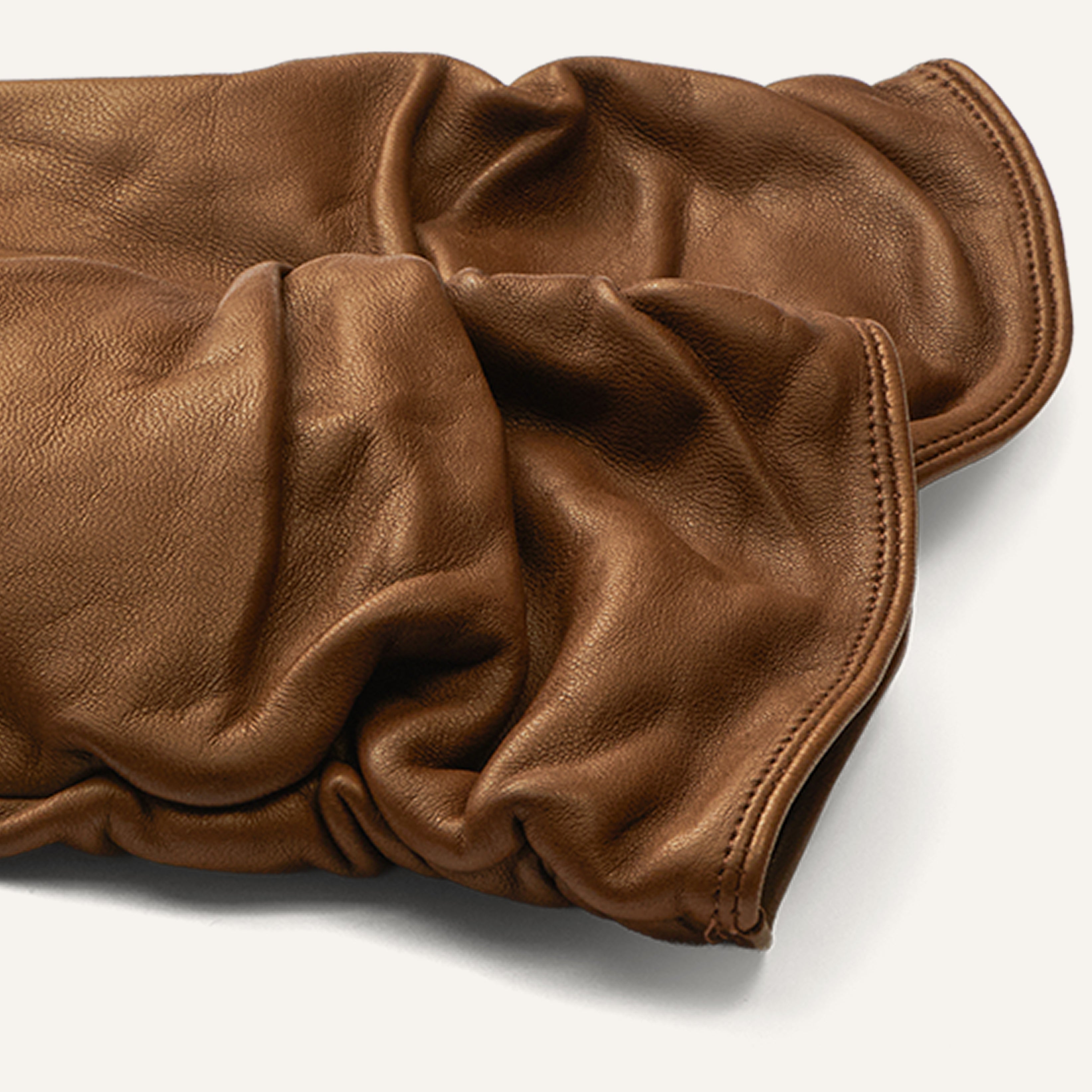 A close-up of two brown leather gloves lying on a white surface, highlighting their soft texture and ruched detailing.