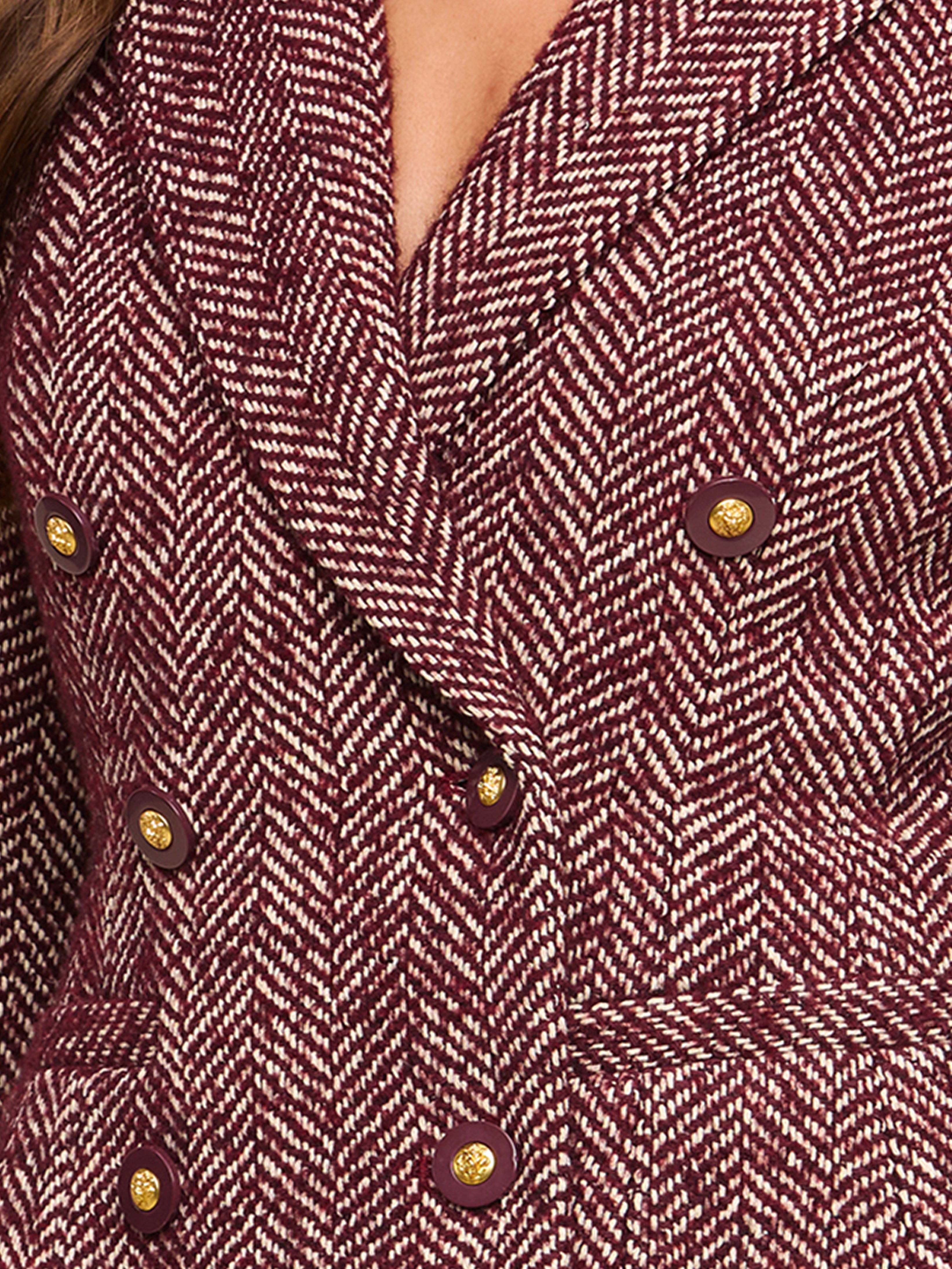 Close-up of a maroon and white herringbone patterned blazer with gold buttons. The double-breasted jacket is being worn by a person, partially visible at the top of the image.