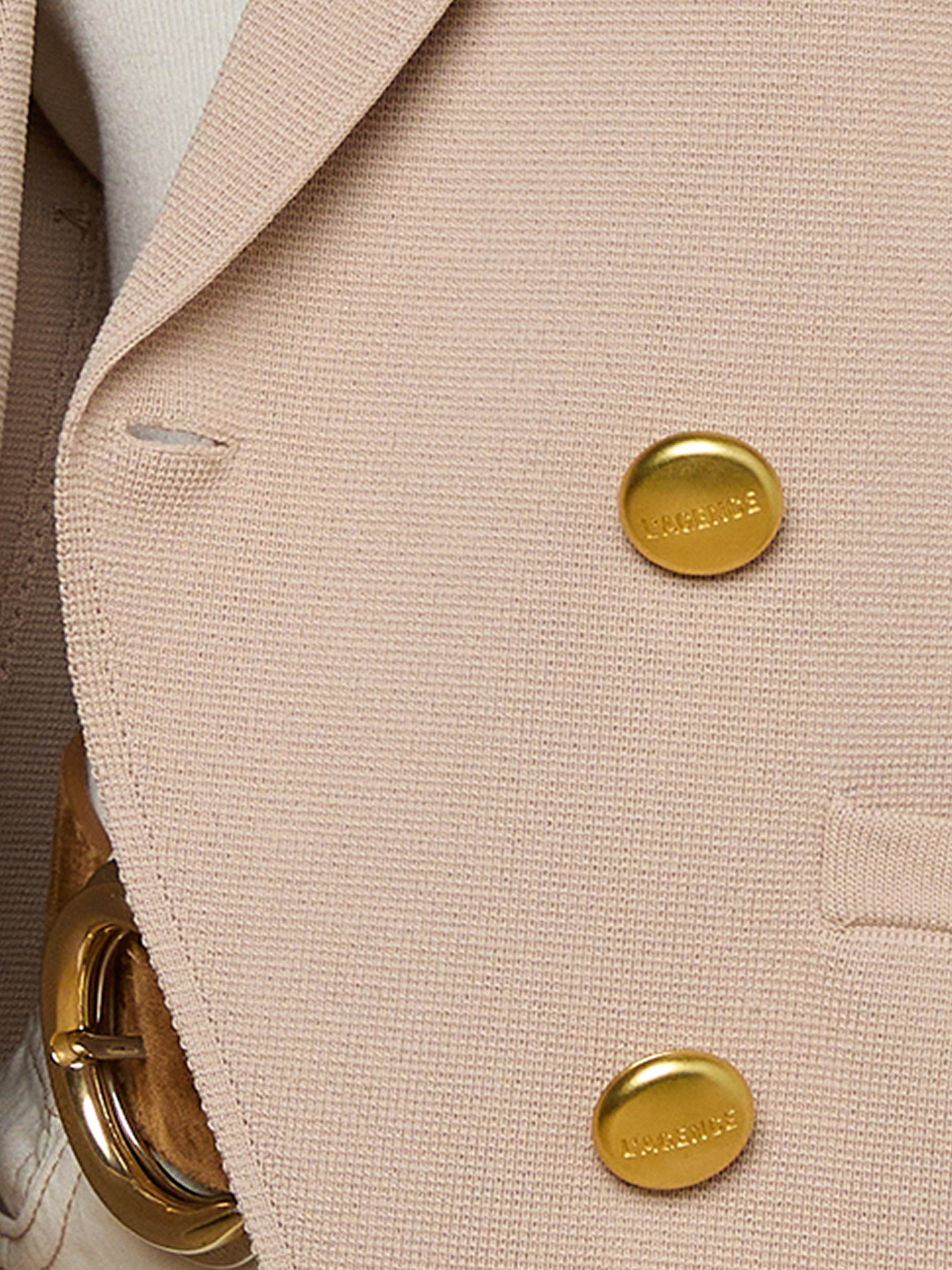 Close-up of a beige textured blazer with two shiny gold buttons and a gold belt buckle partially visible on the left side.