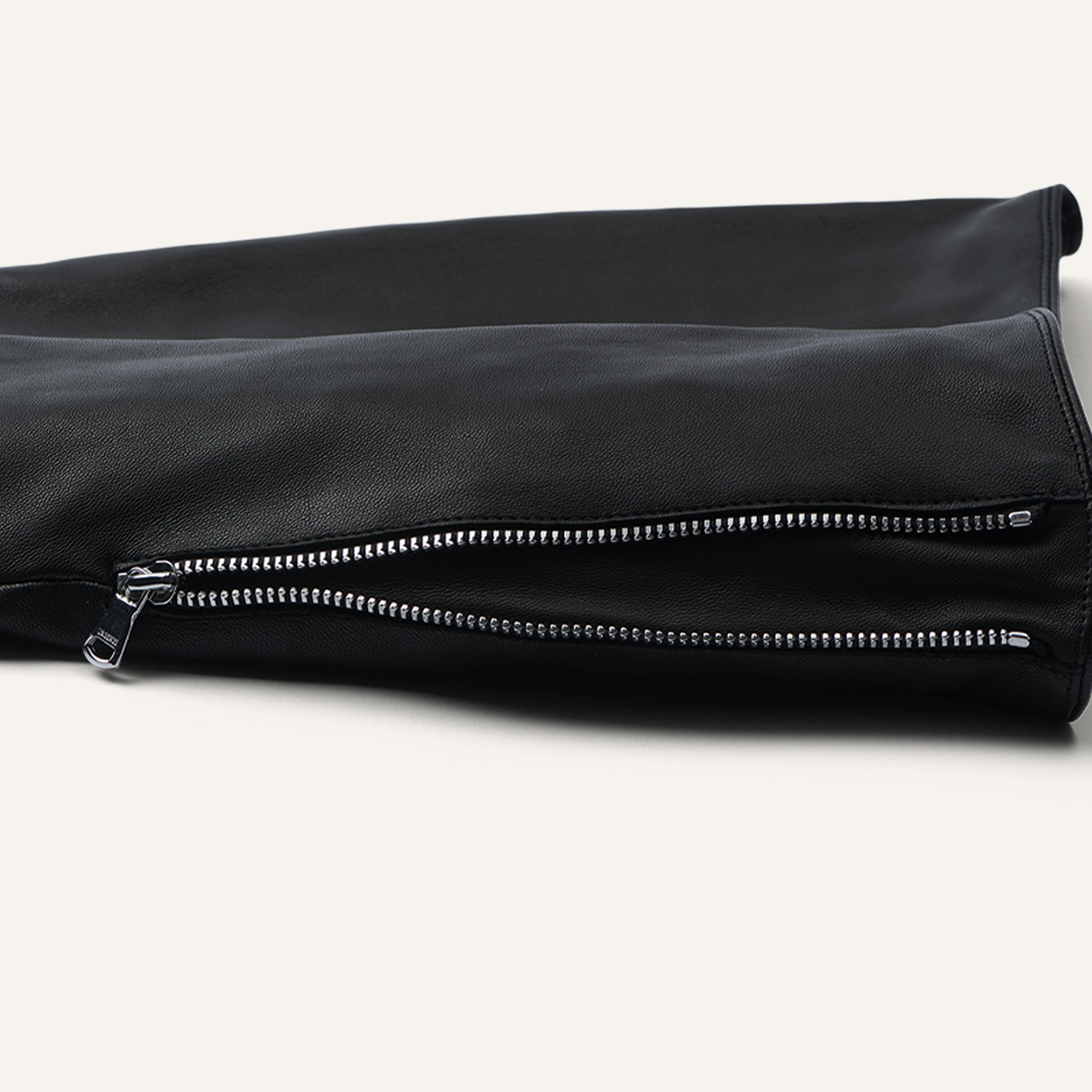 A close-up of a black leather garment showcases two silver zippers running parallel along the fabric, lying flat on a light background.