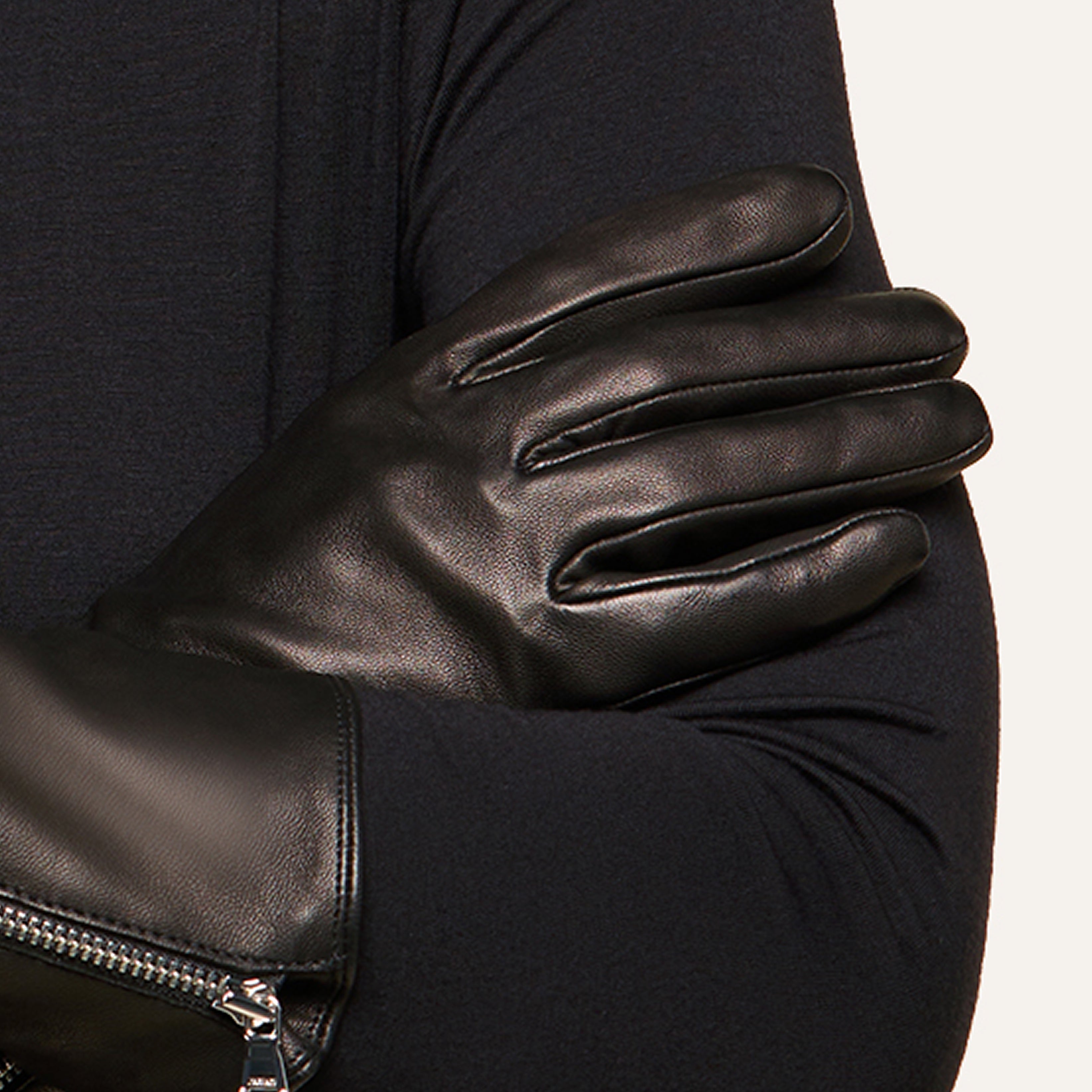 A person wearing shiny black leather gloves crosses their arms over a dark long-sleeve shirt, the supple leather catching the light. Only the torso and arms are visible, with a zipper detail on one glove’s wrist.