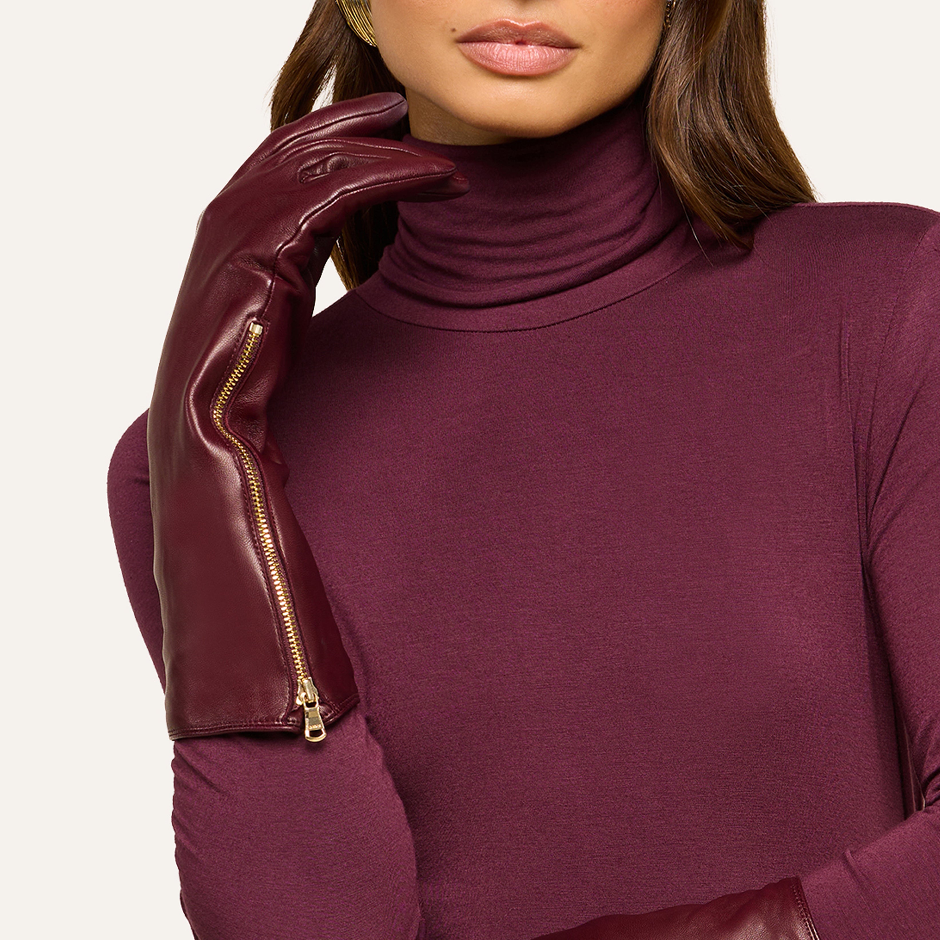Woman in a burgundy turtleneck and matching long leather gloves with gold zippers, posing with one gloved hand near her face. Only the lower half of her face is visible.