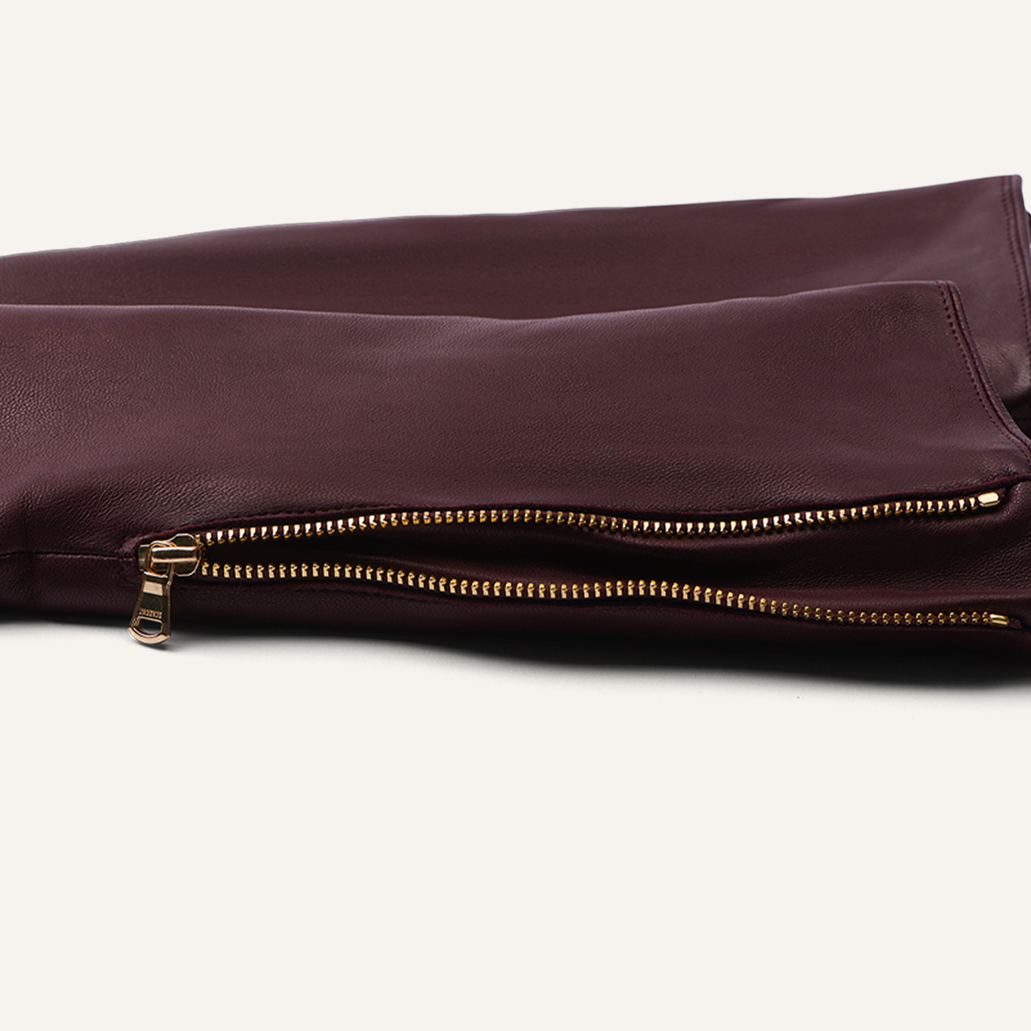 A close-up of a dark burgundy leather sleeve with a gold zipper detail and luxurious texture, laying flat on a light background.