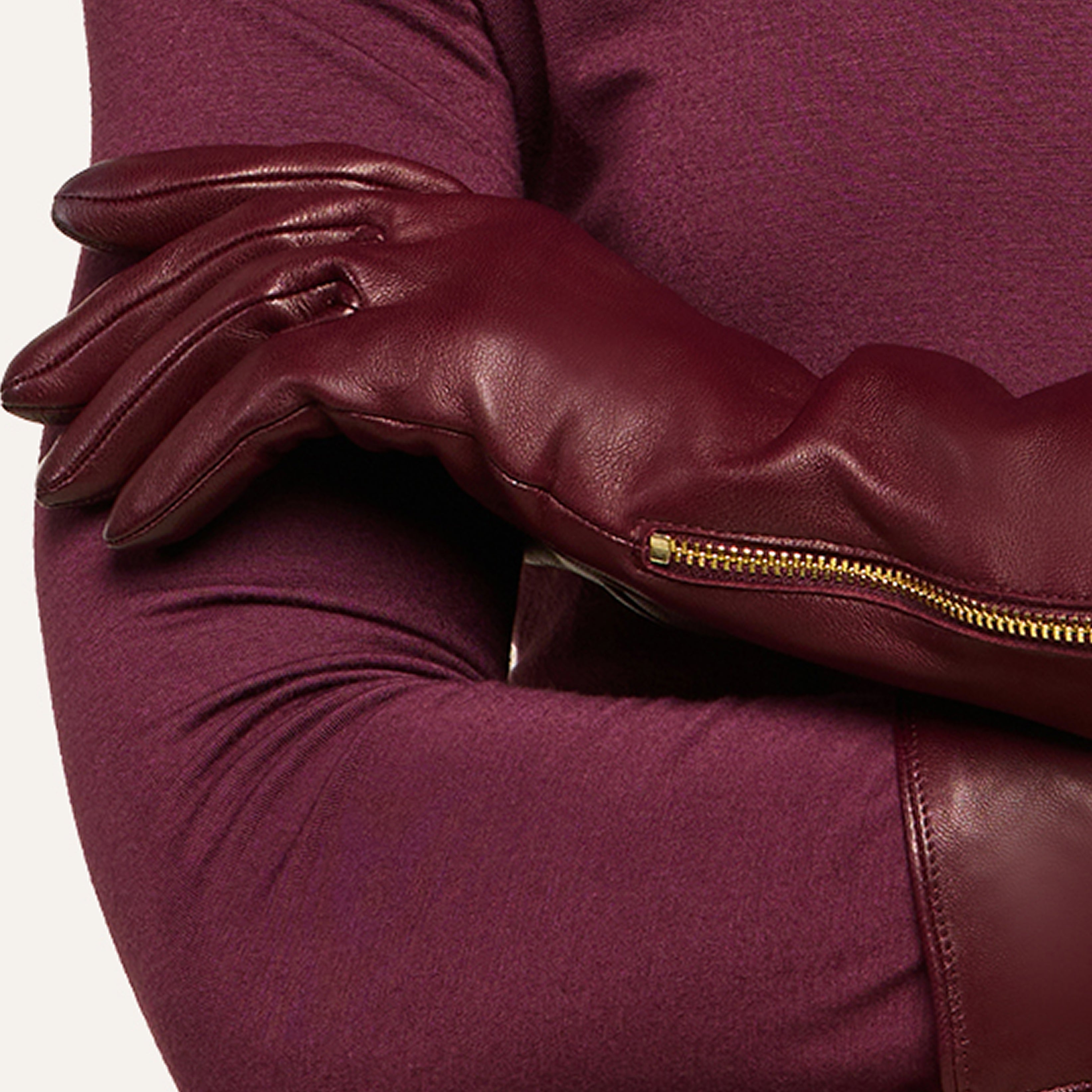 A person wearing a long-sleeve maroon top crosses their arms, displaying burgundy leather gloves accented with a gold zipper detail.