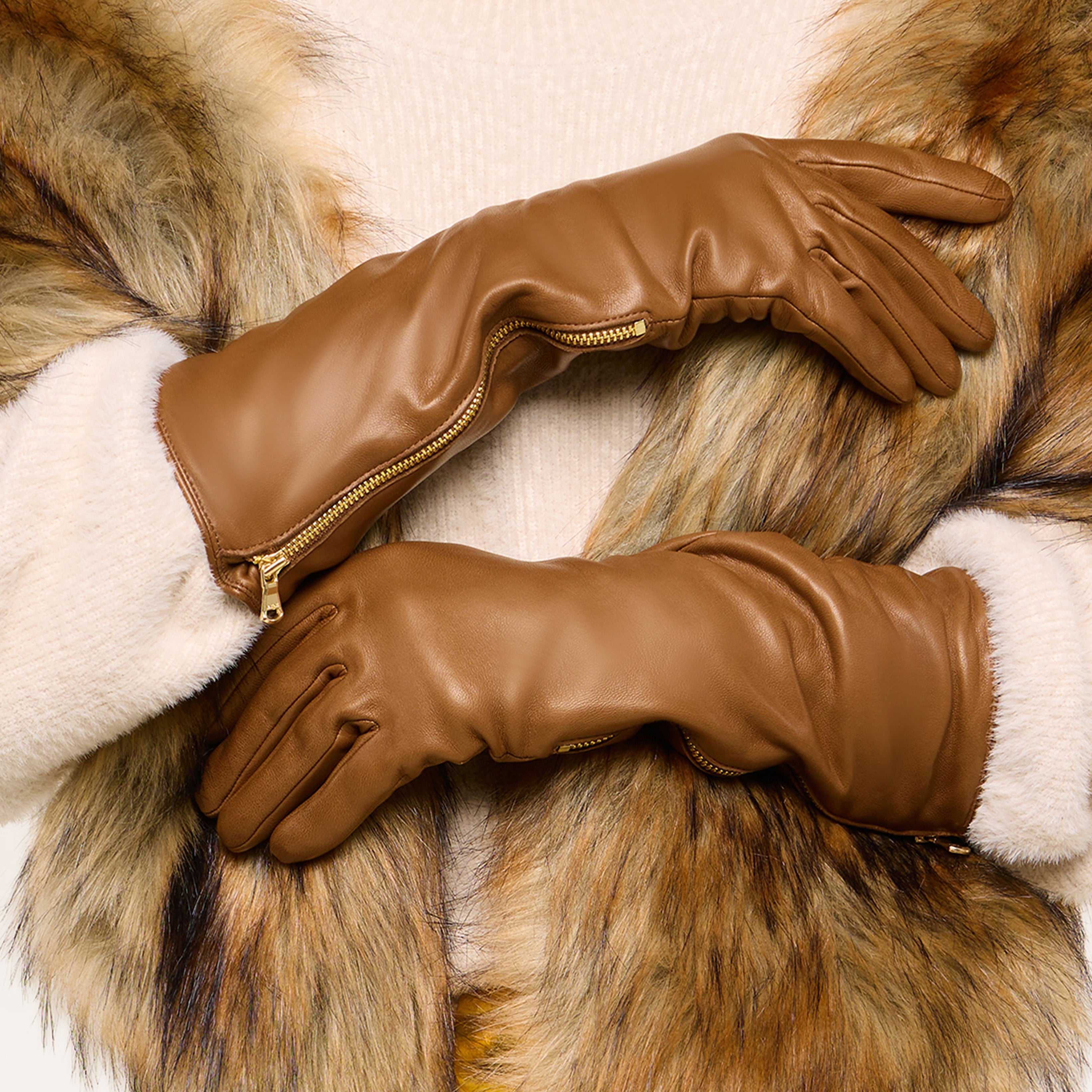 Person wearing a faux fur coat and tan leather gloves with zippers, arms crossed at the wrists, standing against a plain background.