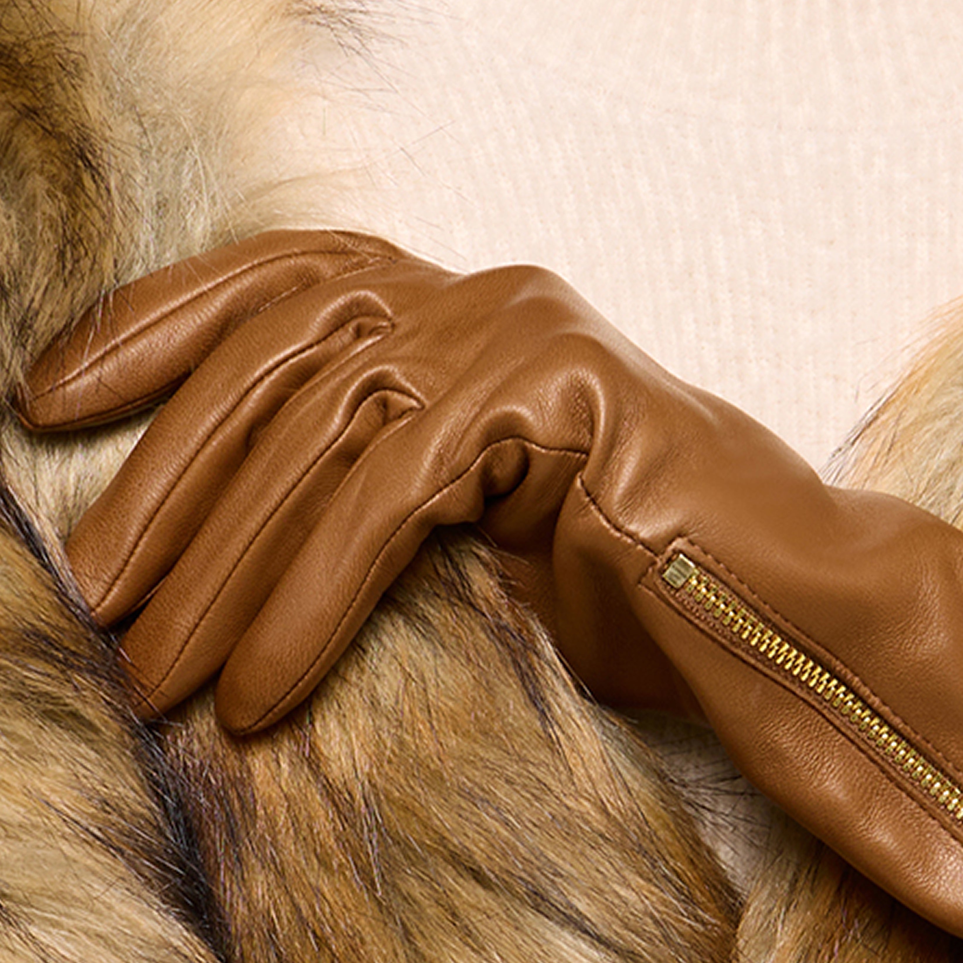 A person wearing a tan leather glove with a zipper rests their hand on a brown fur coat, highlighting the texture of both the glove and the luxurious fur.