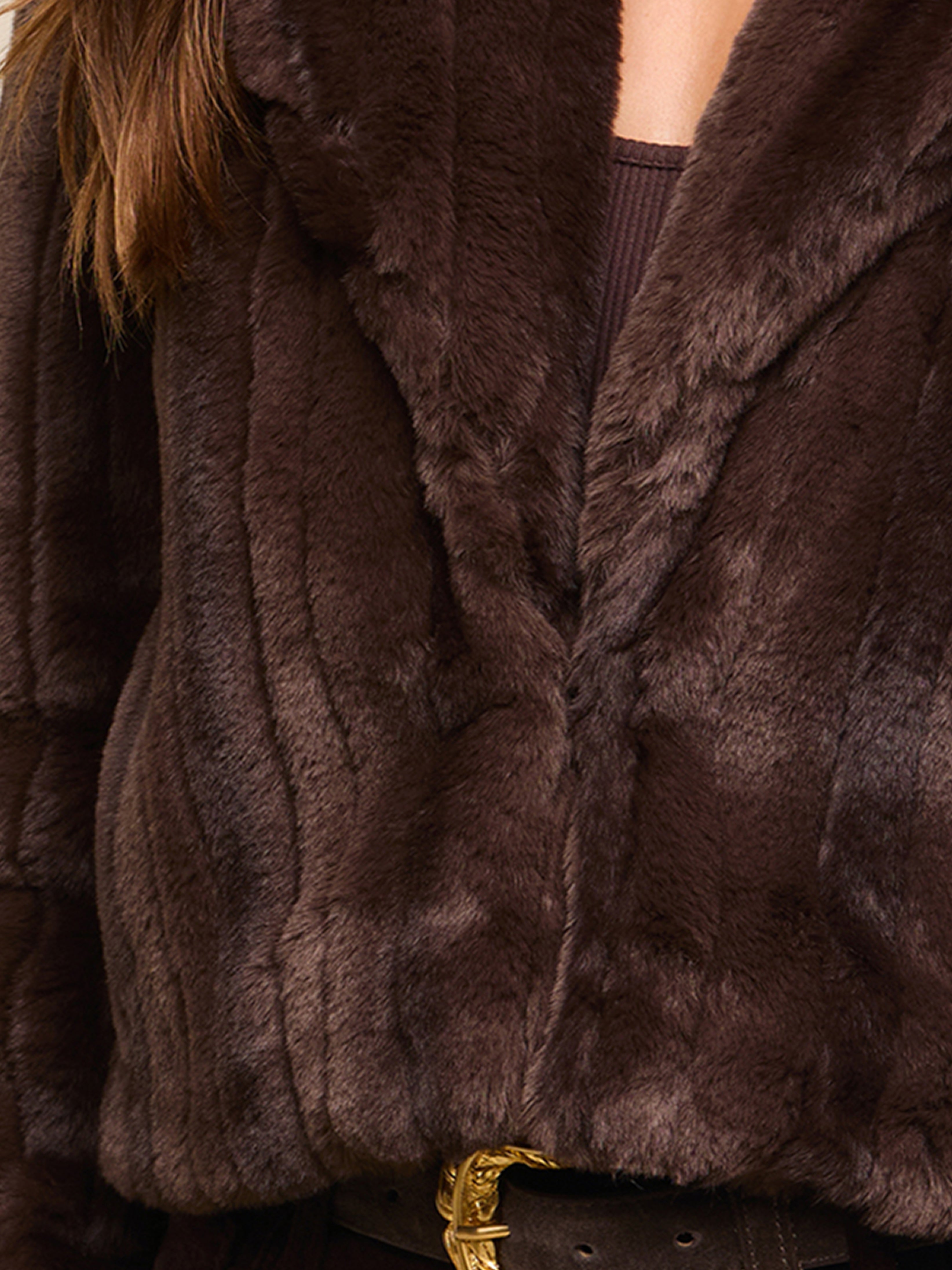 A close-up of a person wearing a dark brown faux fur coat, a brown ribbed top, and a brown belt with a gold buckle. Only the lower face and upper torso are visible.
