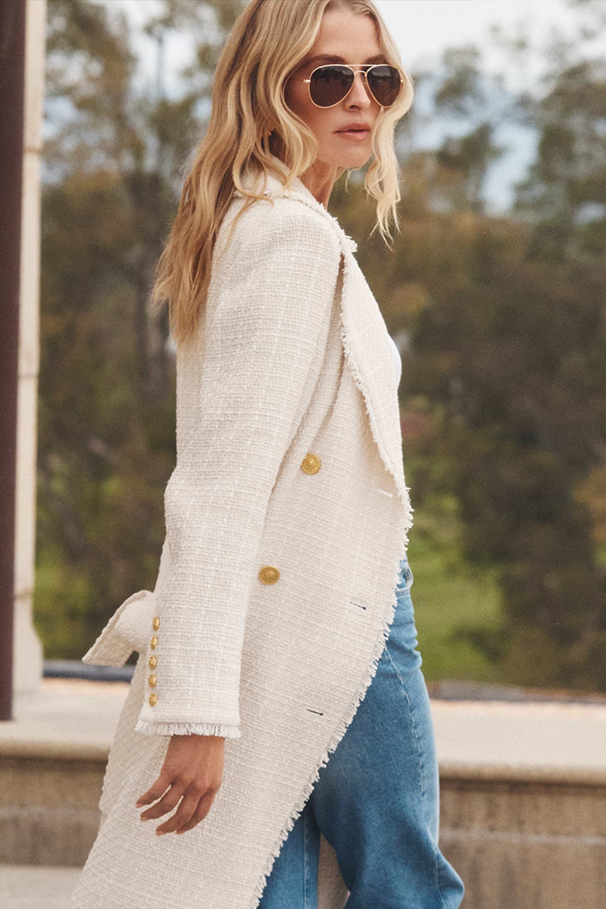 A woman with long blonde hair wearing sunglasses, a textured white coat with gold buttons, and blue jeans walks outdoors by a stone structure, with greenery blurred in the background.