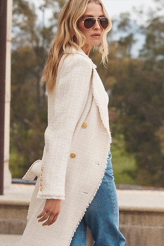 A woman with long blonde hair wearing sunglasses, a textured white coat with gold buttons, and blue jeans walks outdoors by a stone structure, with greenery blurred in the background.