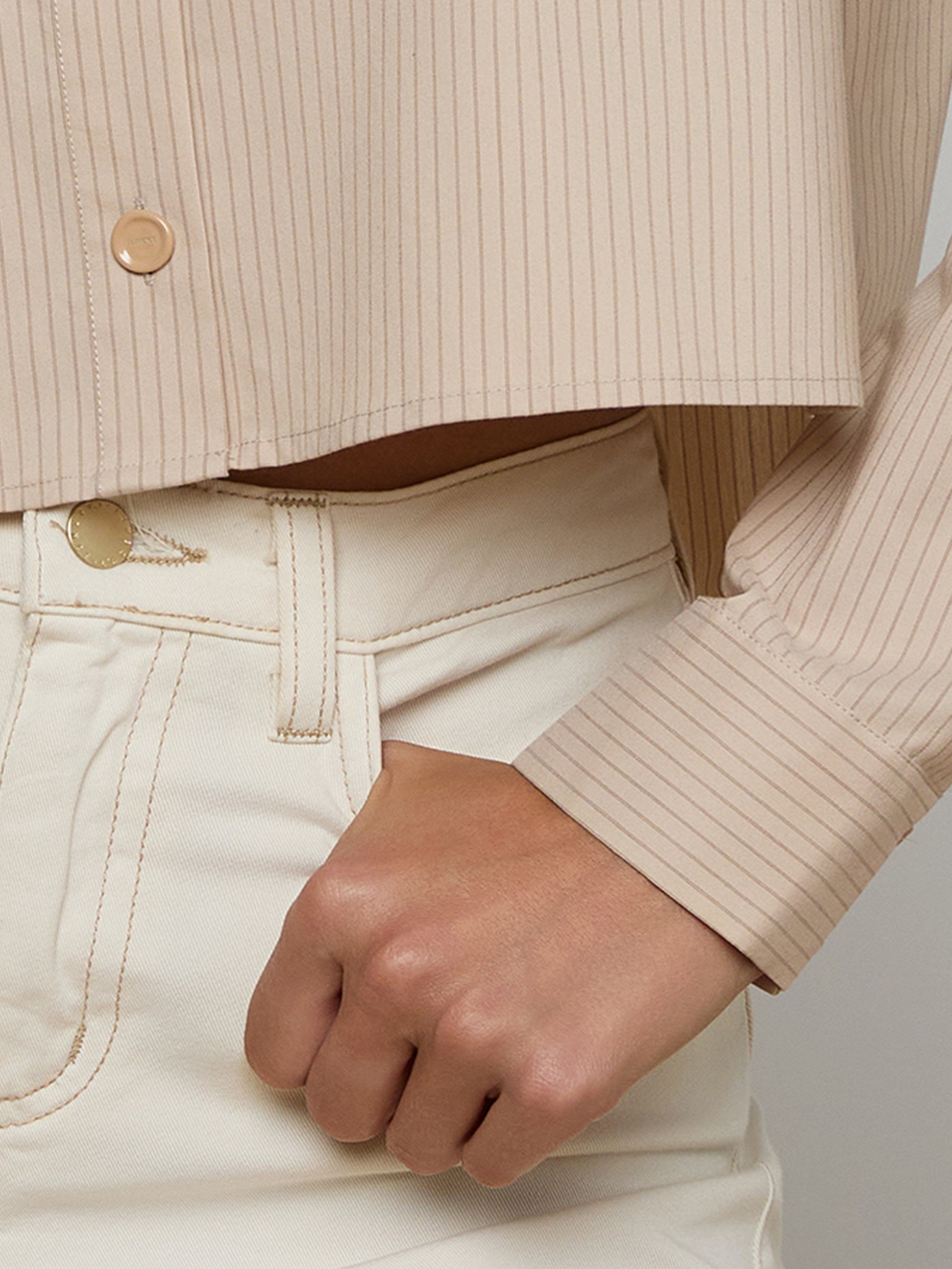 A close-up of a person wearing a beige pinstriped shirt and high-waisted cream pants, with one hand resting in the pants pocket.