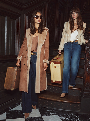 Two women in stylish winter outfits and holding vintage suitcases stand on a dark wood staircase. One wears sunglasses and a tan shearling coat; the other wears blue jeans with the Davy Faux Fur Jacket by L'AGENCE.
