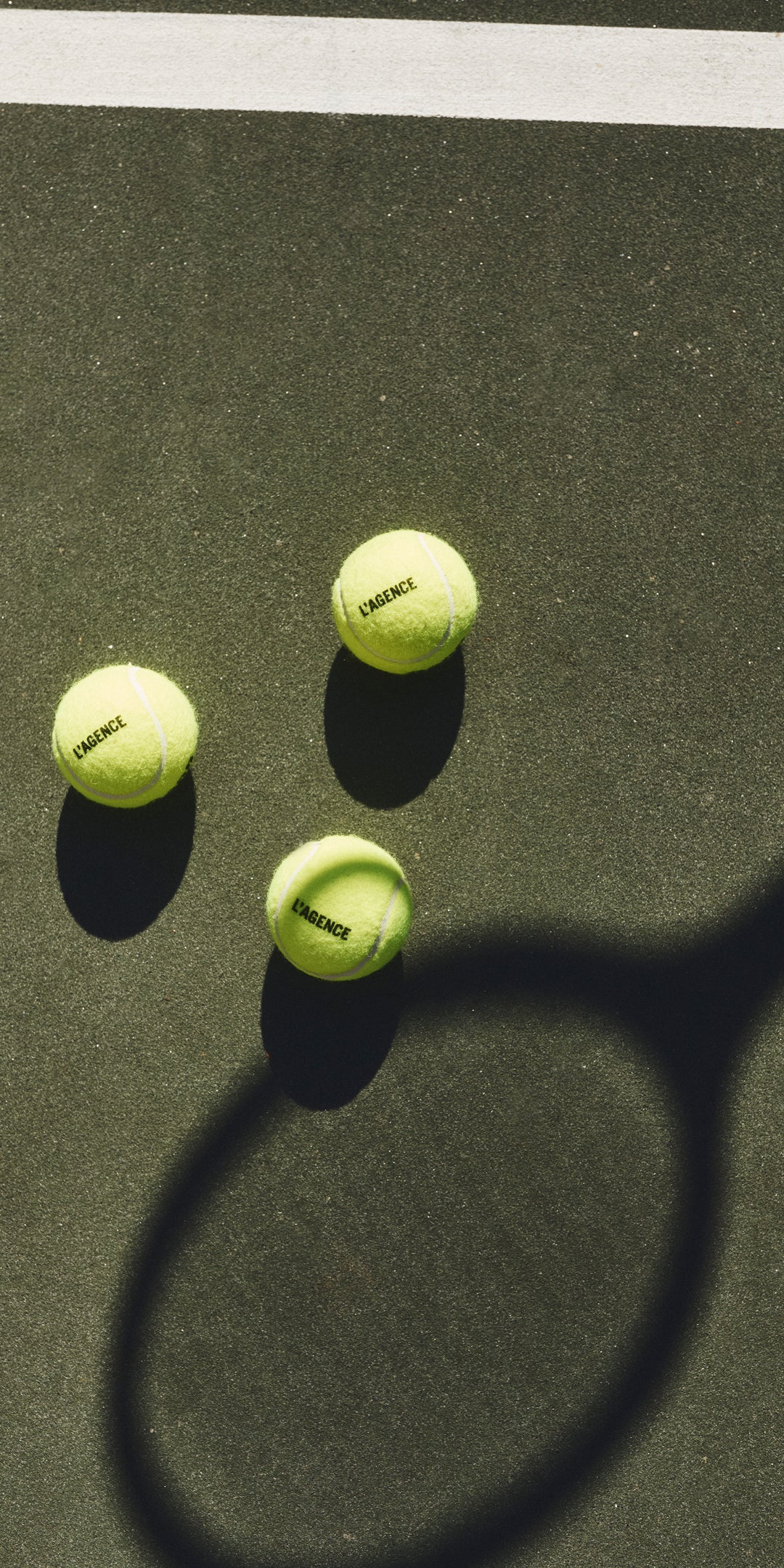 Three yellow tennis balls rest on a tennis court, close to a white line, with the shadow of a tennis racket stretching across the court’s surface.