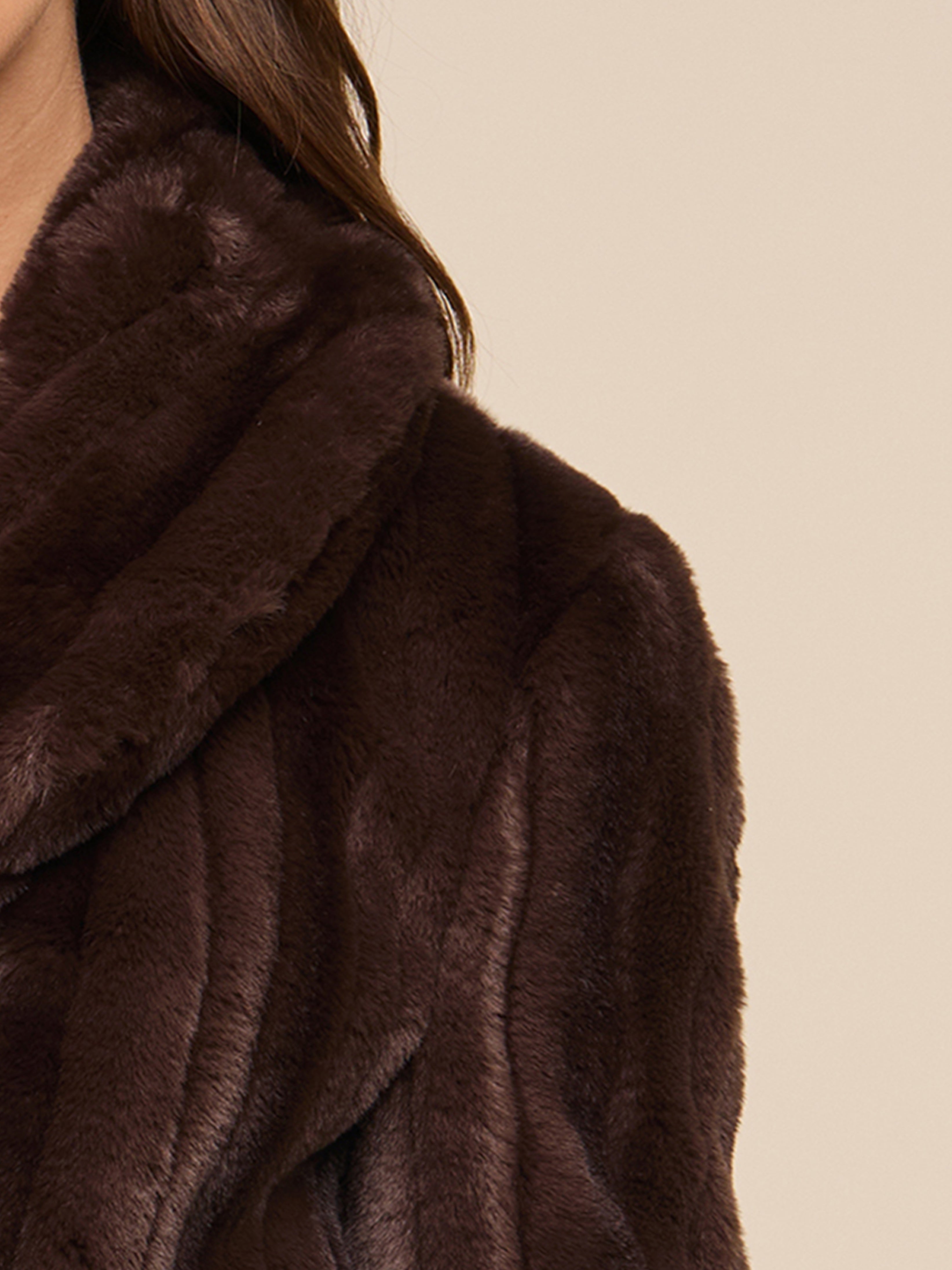 Close-up of a person wearing a dark brown, plush fur coat. Only the shoulder, collar, and part of the persons hair are visible against a beige background.