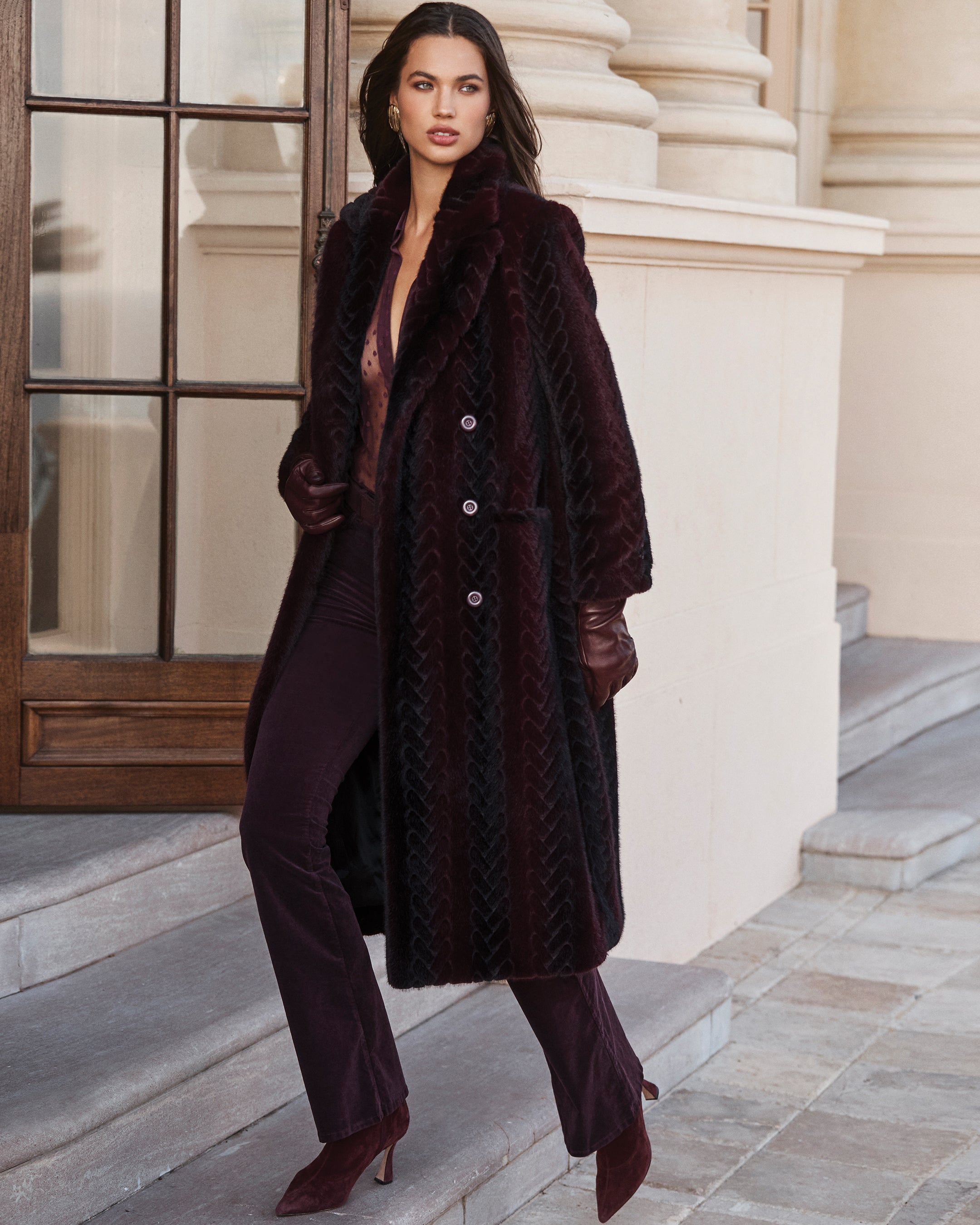 A woman wearing a long, dark textured coat, matching pants, boots, and gloves stands on stone steps outside a building with grand columns and a large window.