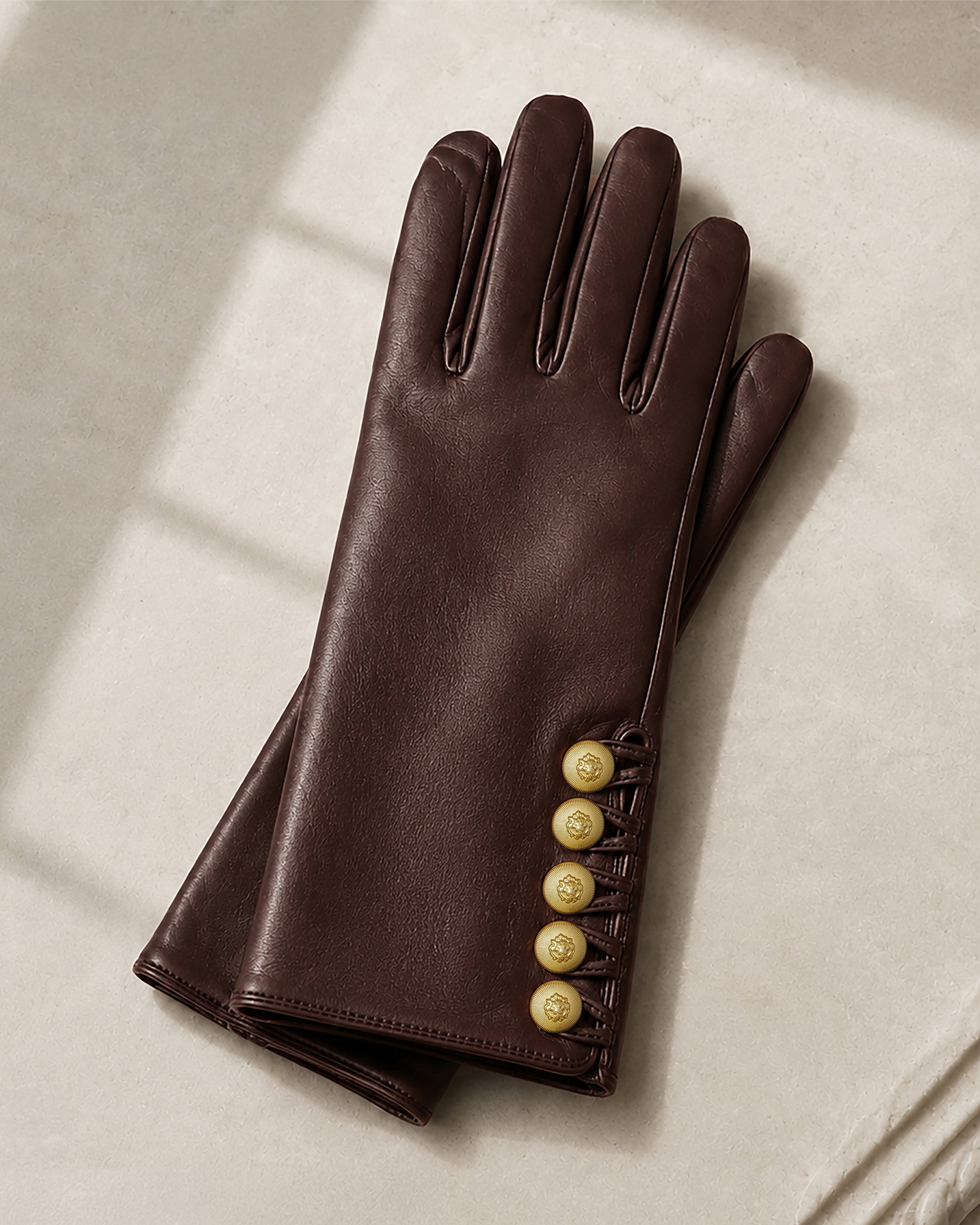 A pair of brown leather gloves with four gold buttons on the wrist, crafted from supple leather and displayed on a light beige surface with soft shadows.