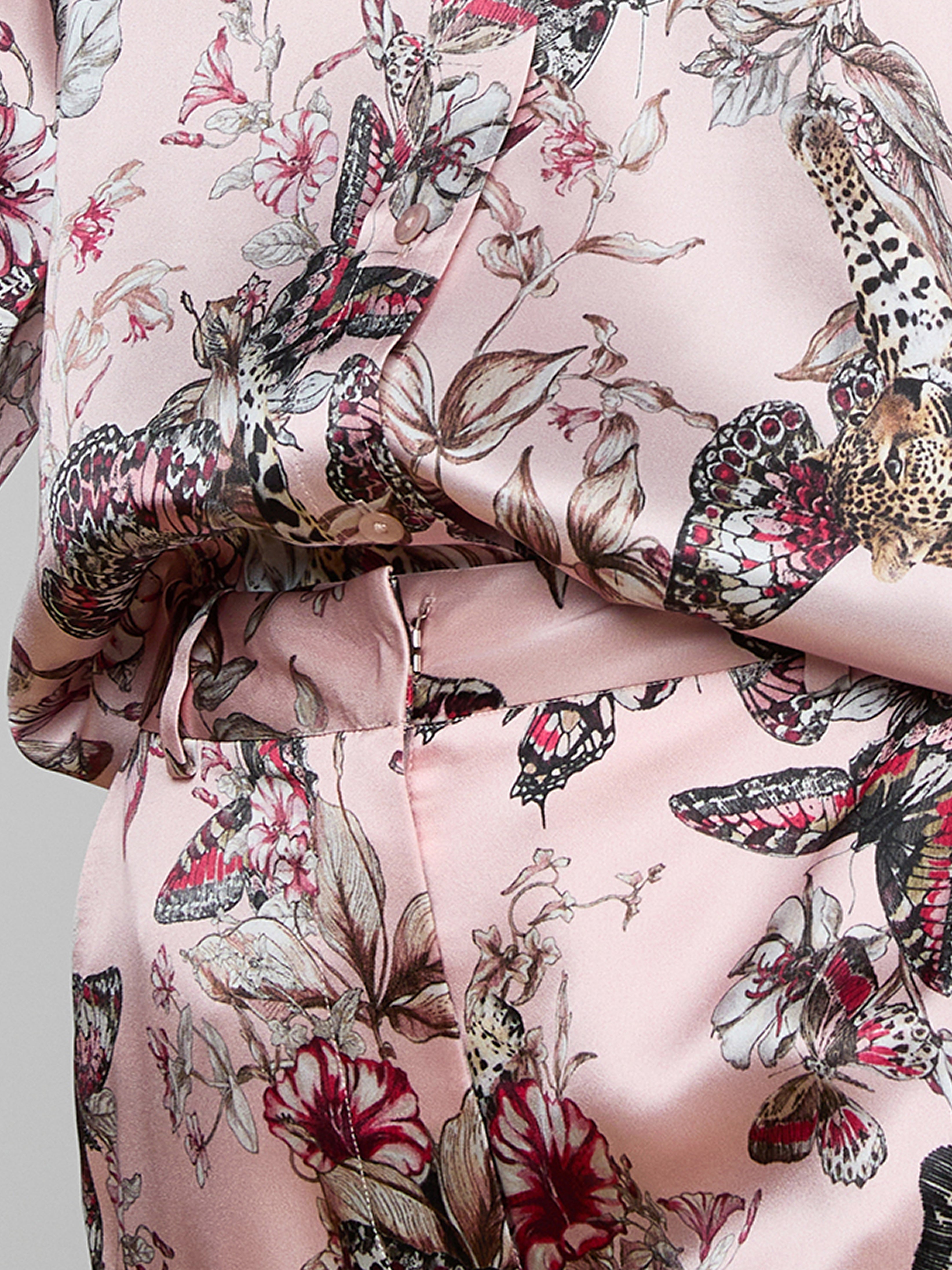 A close-up of a person wearing a light pink outfit with a colorful print featuring butterflies, flowers, and leopard faces. The fabric is silky, showing detailed patterns and a soft sheen.