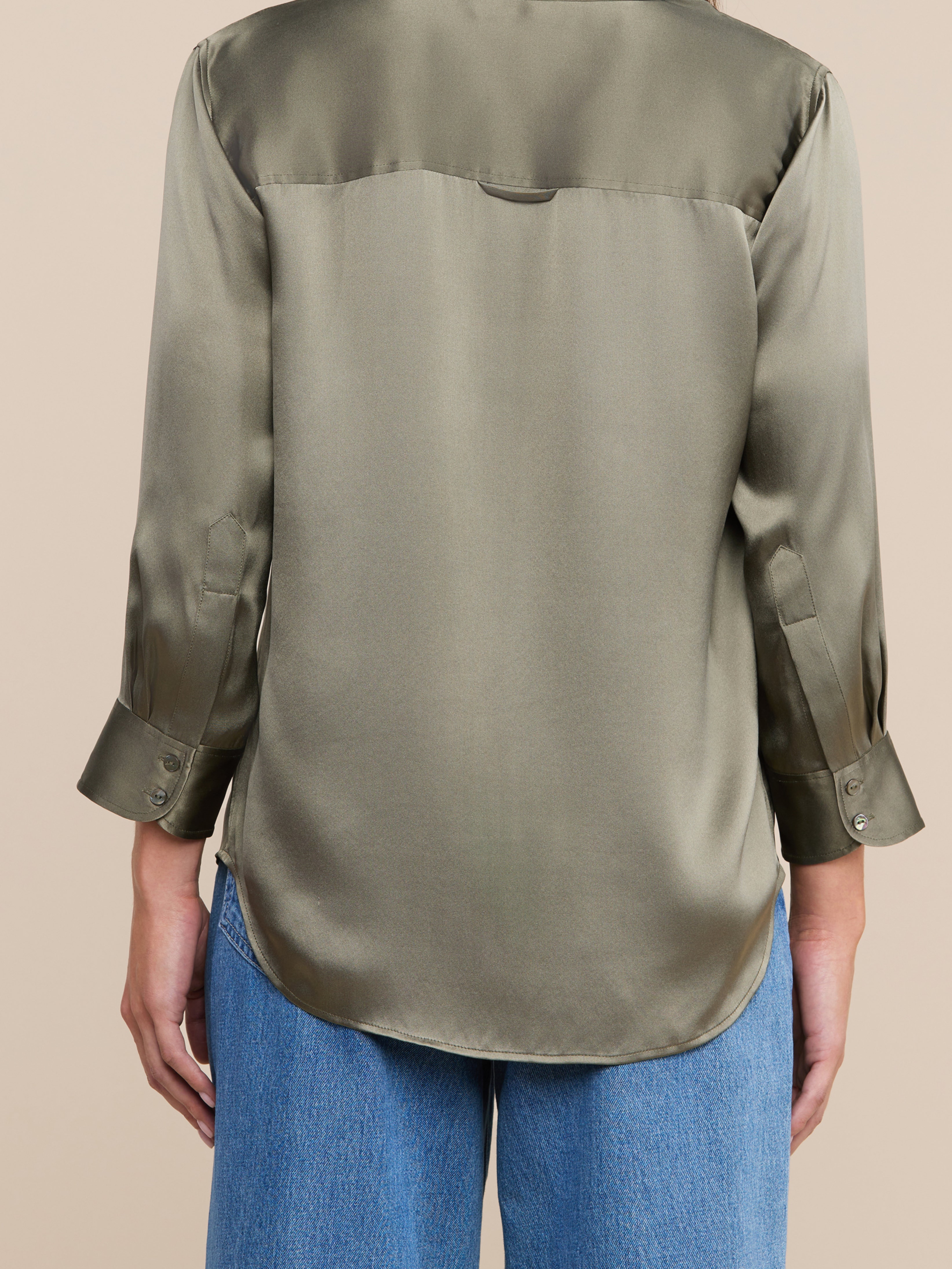 A person wearing a long-sleeved, olive green satin blouse and blue jeans is shown from the back against a neutral background. The blouse has a curved hem and buttoned cuffs.