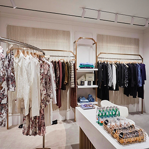 Clothing boutique with racks of patterned and solid garments, shelves with folded clothes, and a table displaying bottled water and pastries on trays in the foreground.