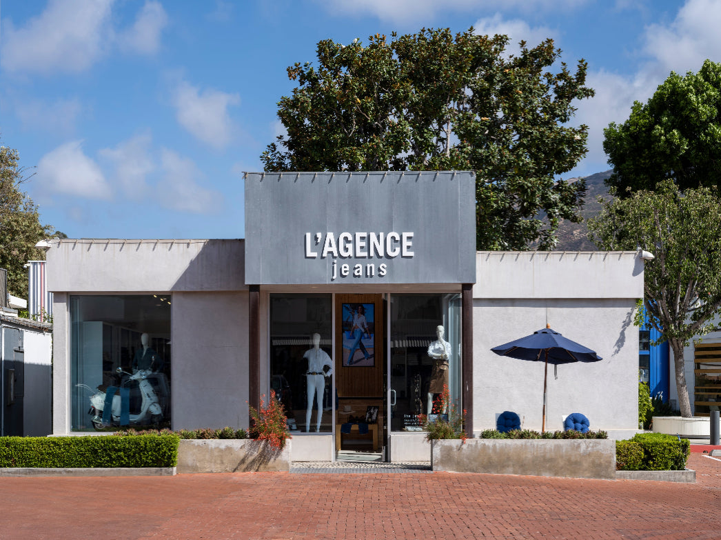 LAGENCE jeans storefront with mannequins in window displays, a dark umbrella on the right, and trees in the background under a blue sky with clouds.