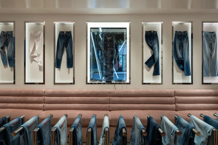 A clothing store display with jeans and shirts hanging in lighted wall frames, a cushioned bench below, and more jeans neatly arranged on racks in the foreground.