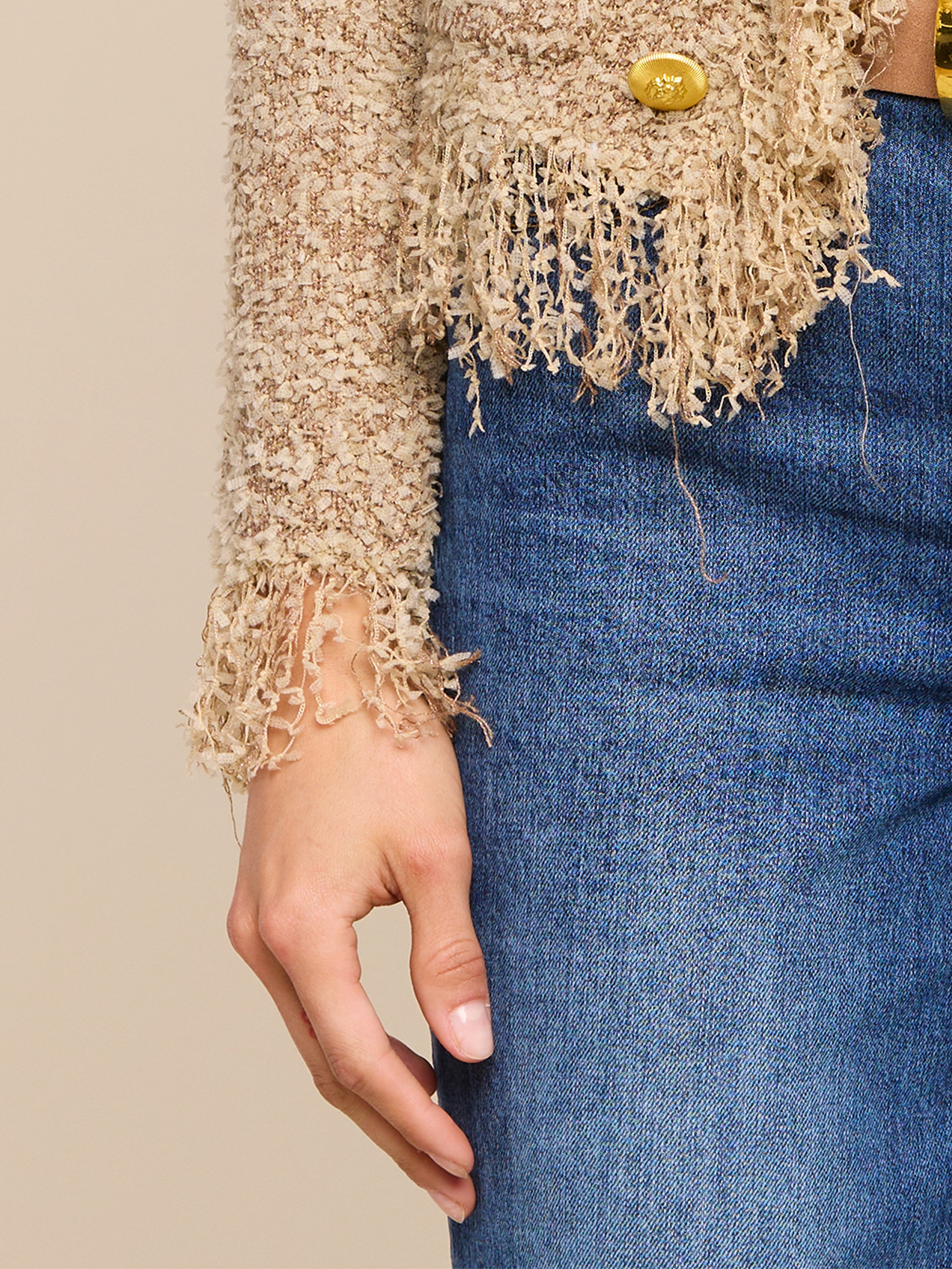 A person wears blue jeans and a textured beige jacket with frayed edges on the sleeve and hem. Only the lower arm and part of the torso are visible.