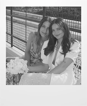 Two women sit closely together outdoors, smiling at the camera. One wears a floral dress and the other a white outfit while holding a bouquet of flowers. They are next to a fence and the image is in black and white.