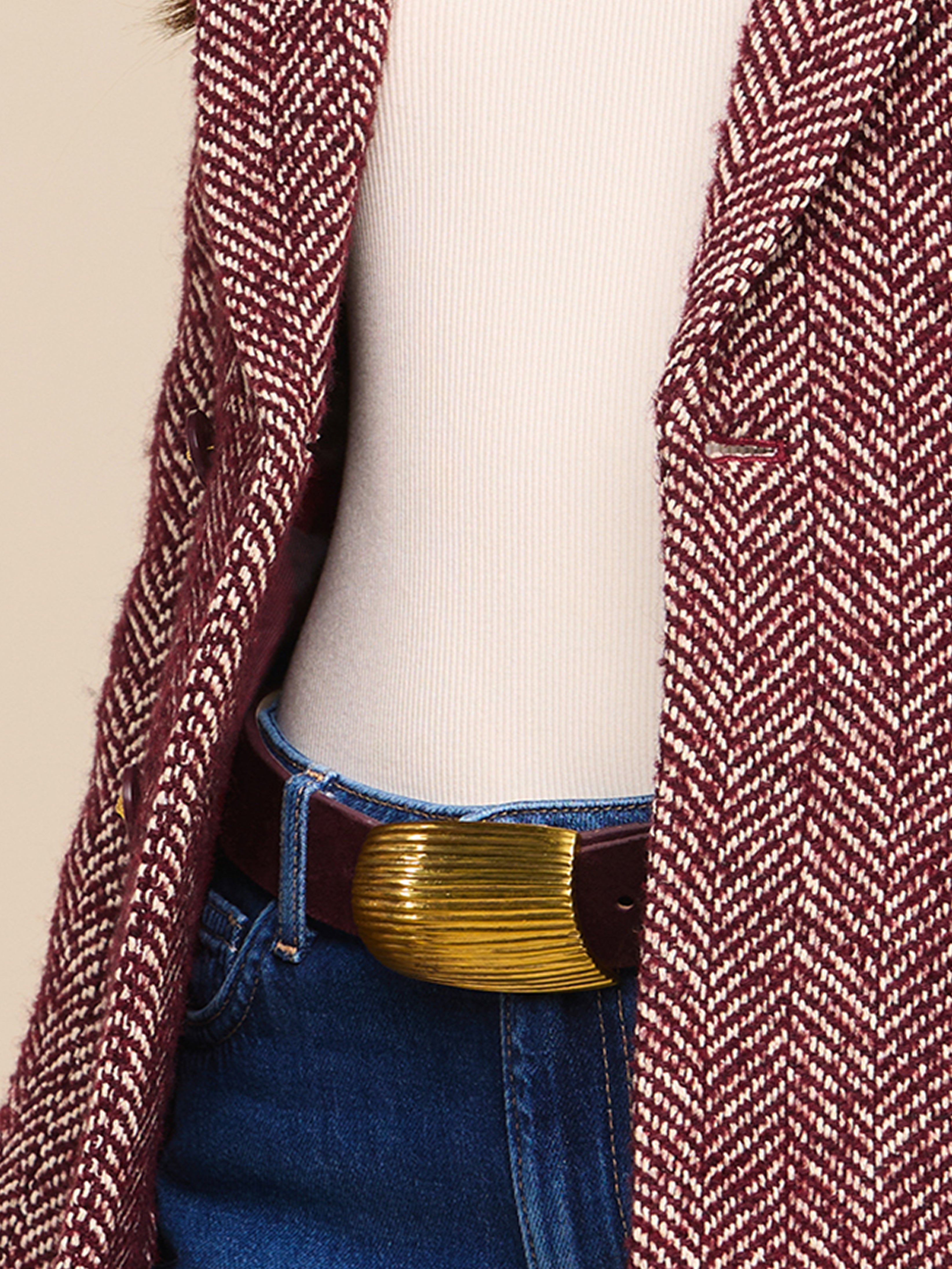 Close-up of a person wearing a maroon and white herringbone coat, a cream ribbed top, blue jeans, and a brown belt with a large gold buckle.