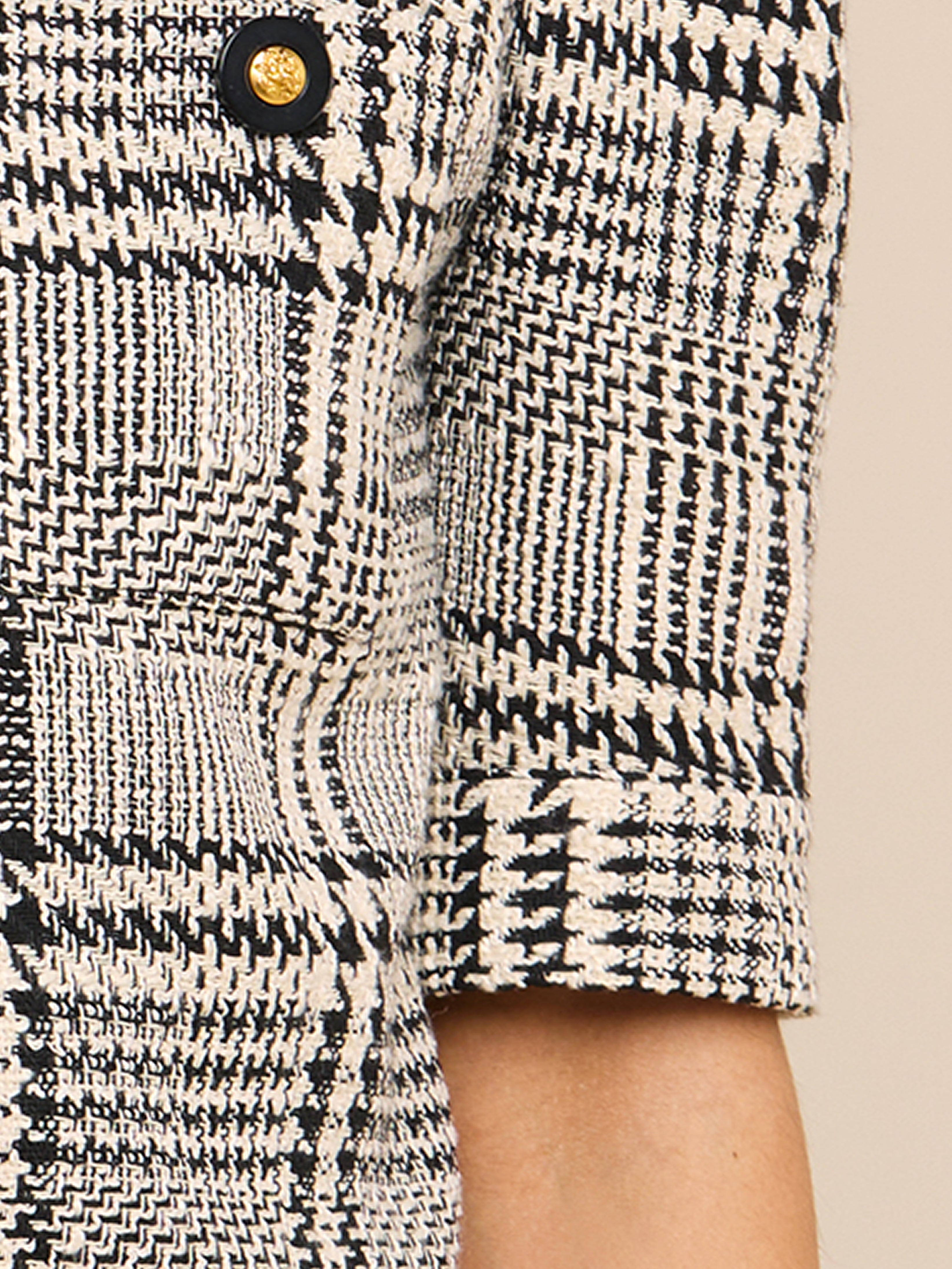 Close-up of a black and white houndstooth-patterned blazer sleeve with a visible gold and black button. The fabric texture and details of the jacket design are clearly shown.