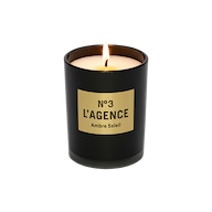 A lit, hand-poured Ambre Soleil candle in a black glass holder with a gold label sits elegantly on a plain white background. Crafted from carbon neutral soy-blend wax, this candle promises a long burn time while adding warmth and sophistication to any space.