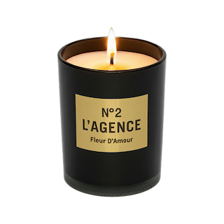 A lit Fleur D’Amour hand-poured candle in a black glass jar with a beige rectangular label is displayed against a white background, featuring a luxurious baccara rose scent.