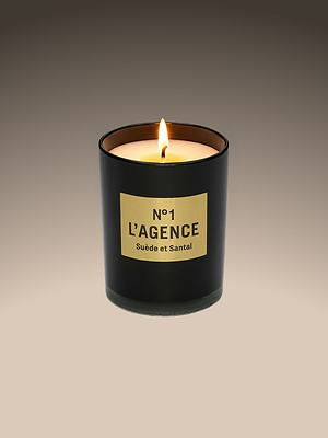 A lit Suède et Santal candle rests in a sleek black glass holder with a beige label, crafted from soy-blend wax and set against a soft gradient background.