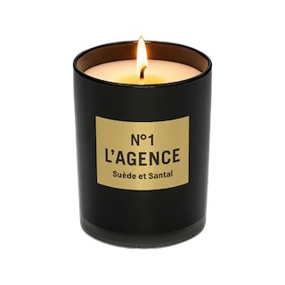 A lit Suède et Santal soy-blend candle in a sleek black glass holder with a beige label sits elegantly against a plain white background, capturing the sophisticated essence of Southern California.